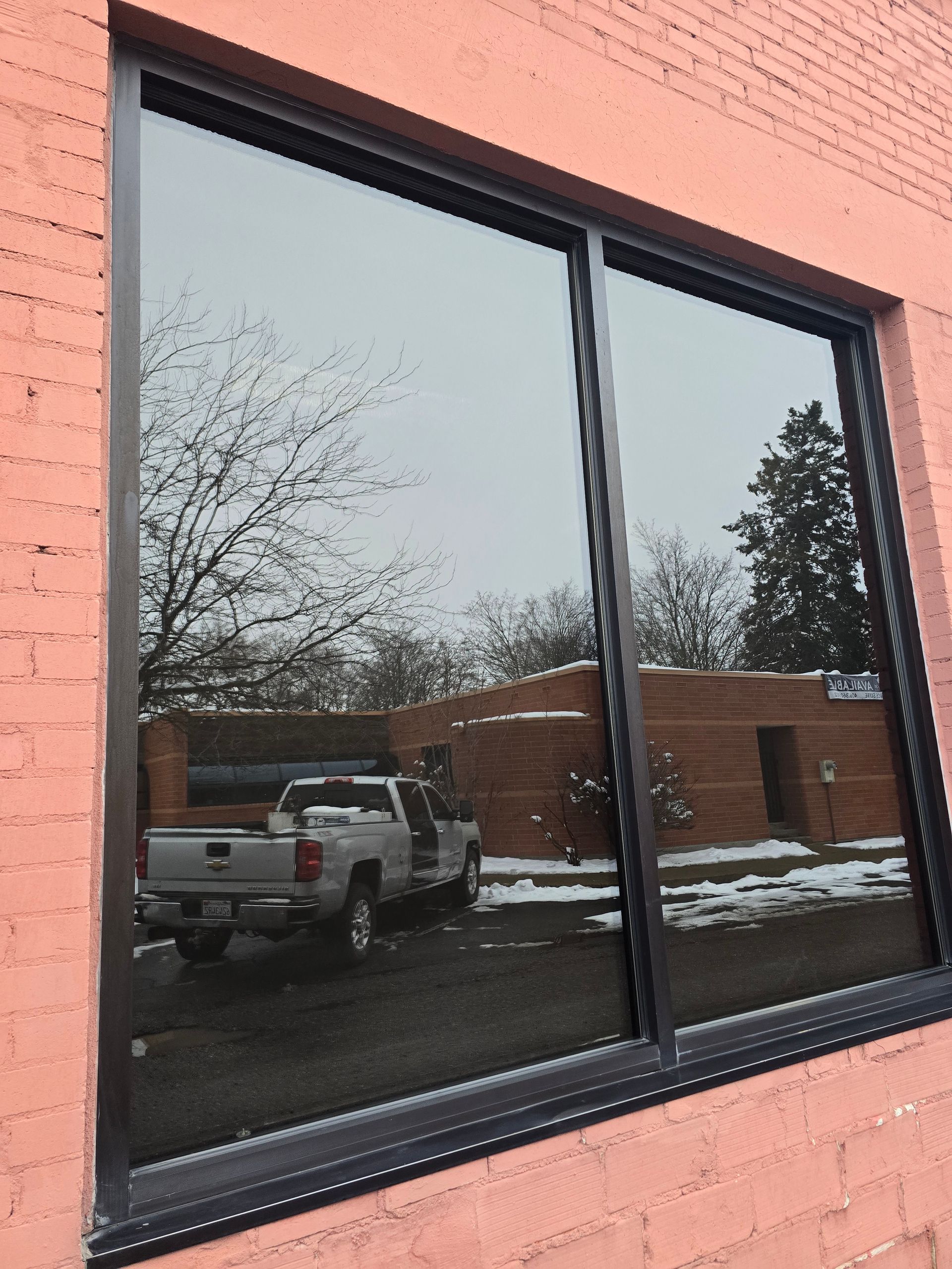 A dark-framed window on a pink brick building reflects a parking lot, trees, and a silver pickup truck.