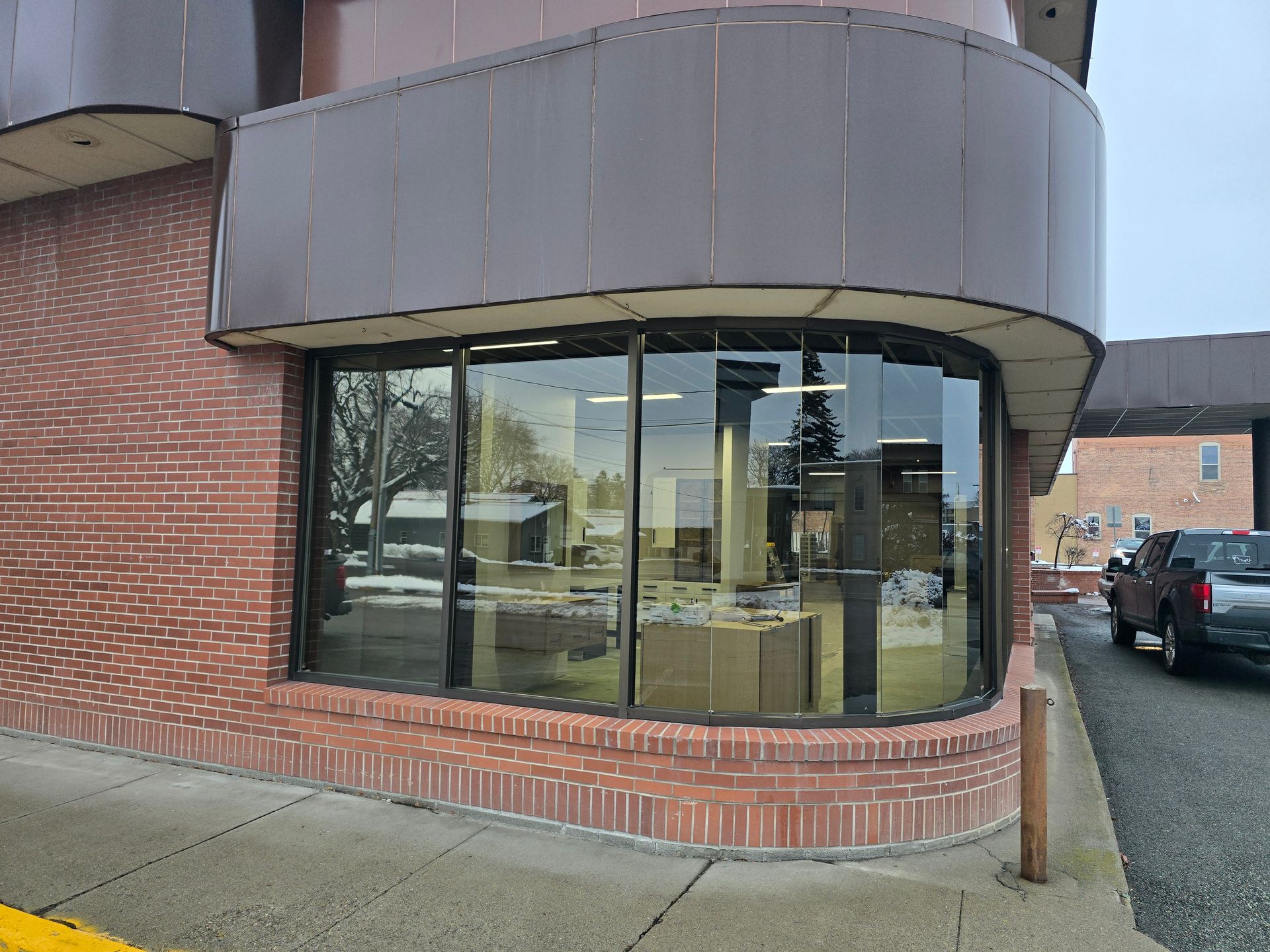 A curved brick building corner with a large glass storefront window, dark metal trim, and a truck parked nearby.