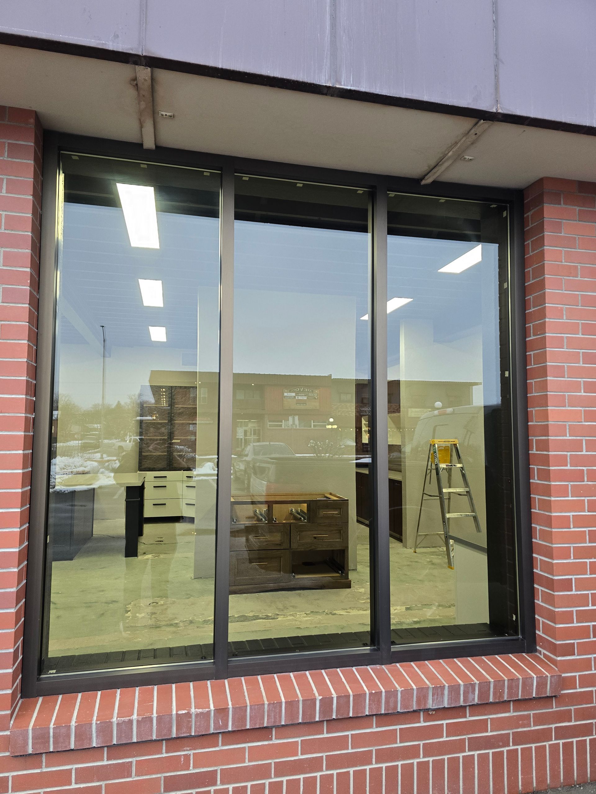 A large three-pane storefront window set in a red brick wall, showing an interior space with a ladder and furniture.
