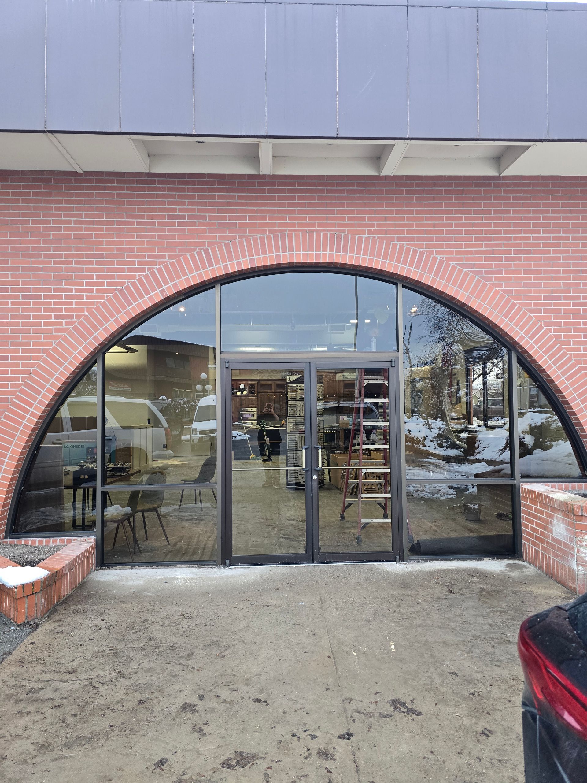 A brick building entrance featuring a large, arched glass door and window display reflecting a snowy parking lot.