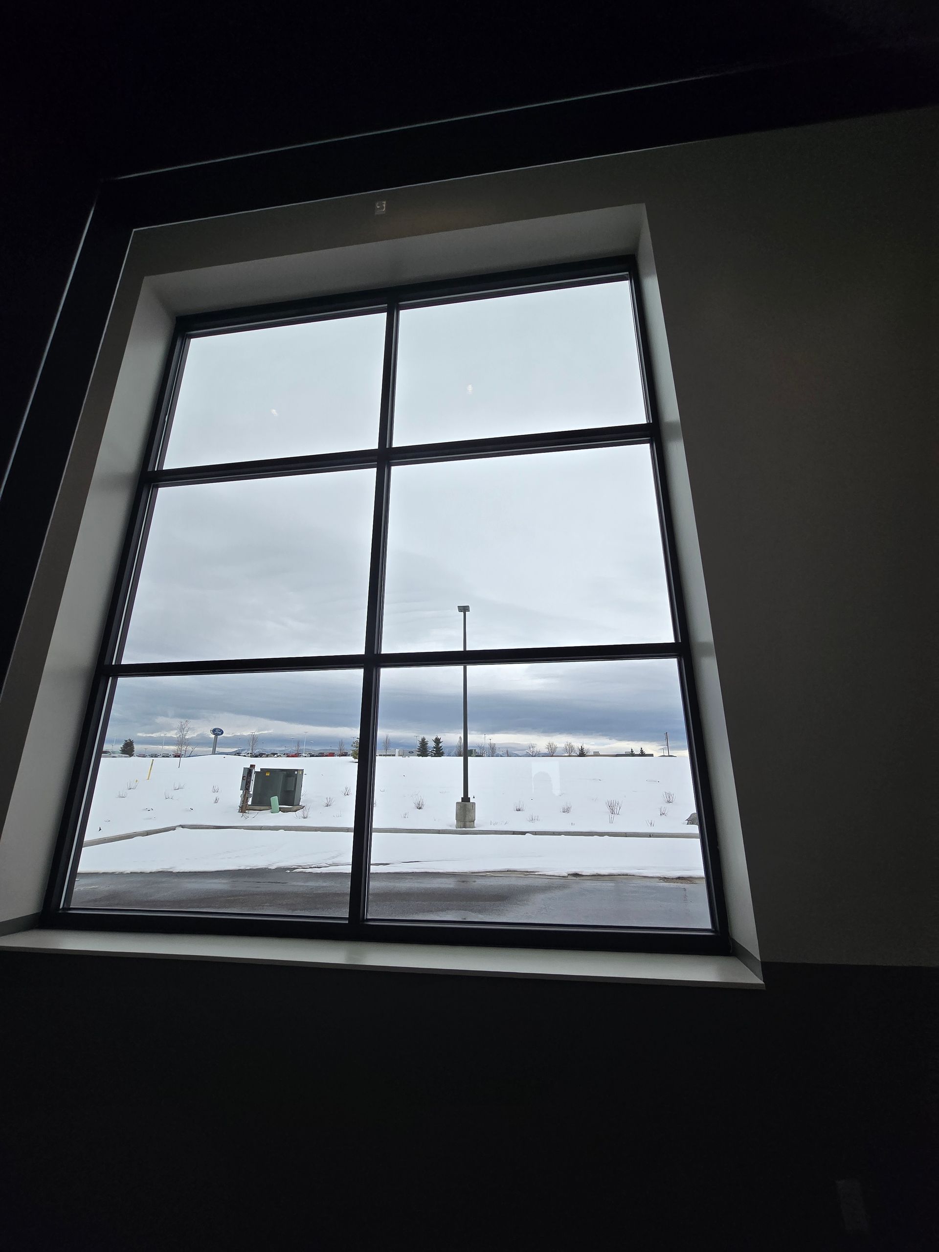 A window view of a snowy, overcast parking lot with a single light pole, framed by dark interior walls.