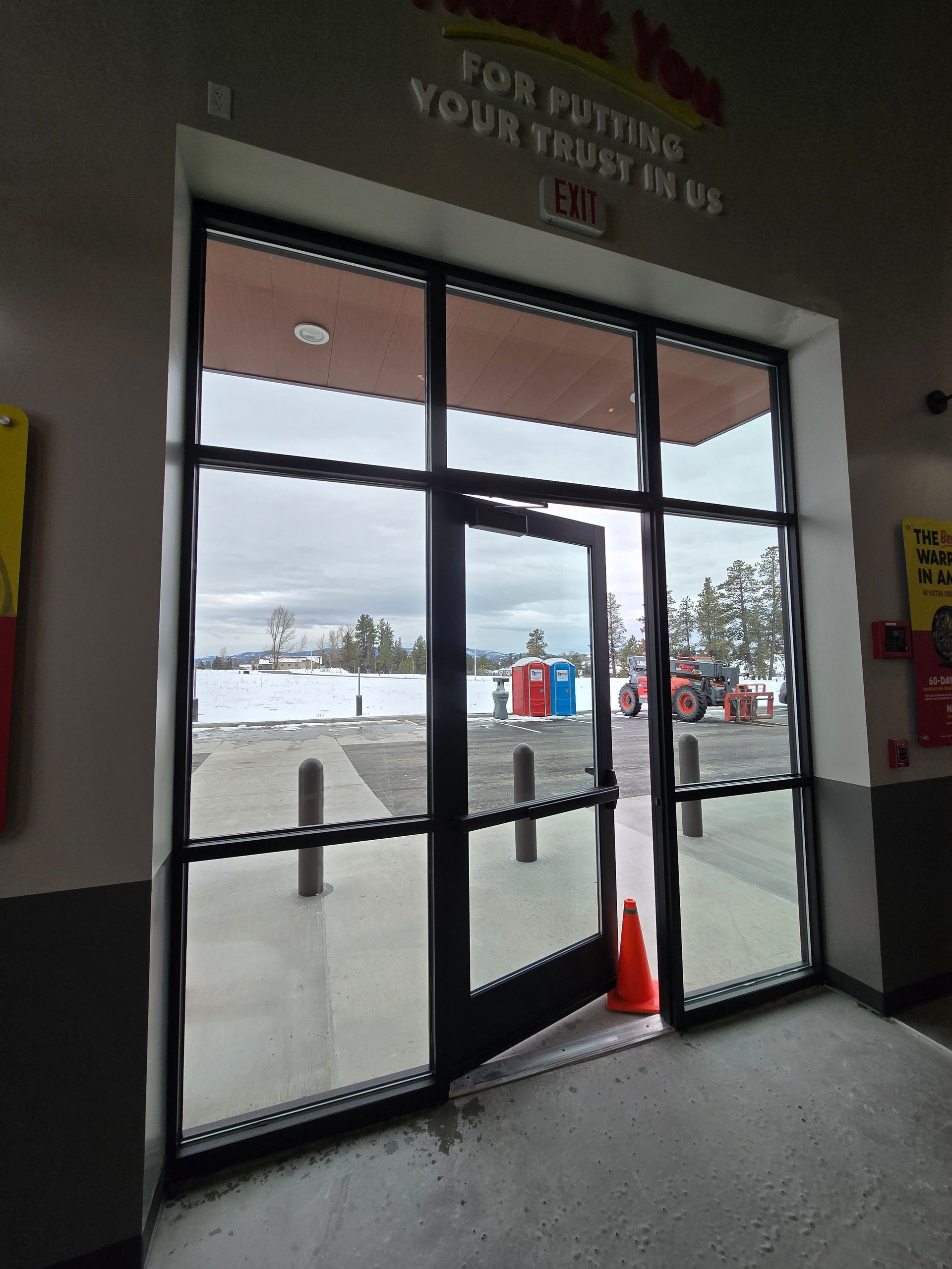 A broken commercial door in a building entrance, propped open with an orange traffic cone, showing an outdoor parking lot.