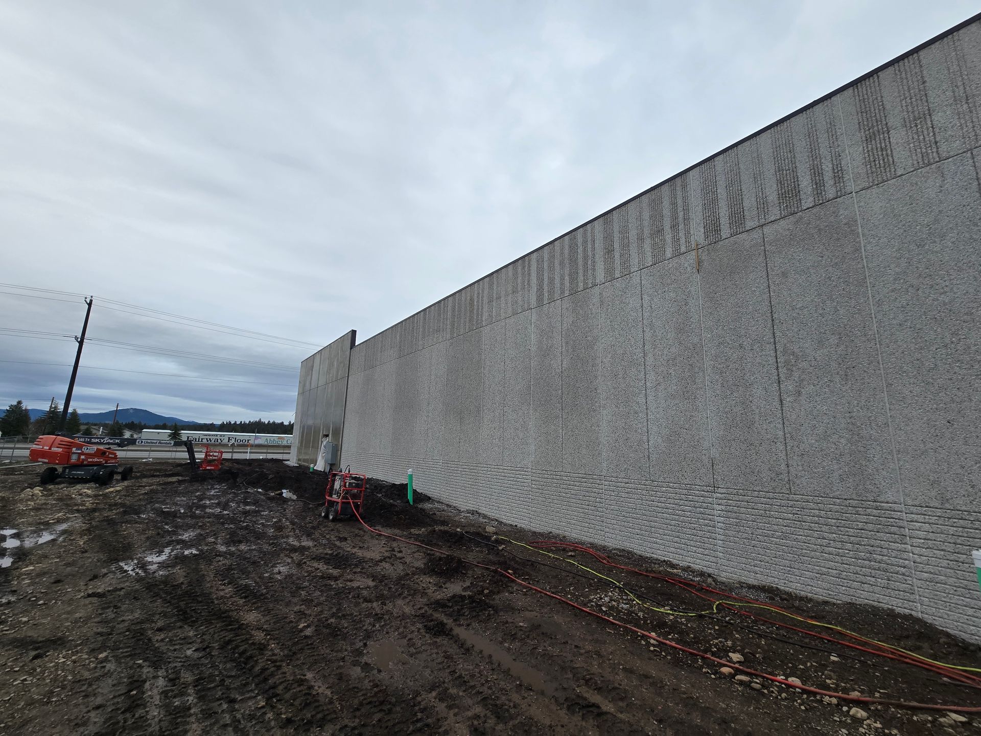 A tall, textured gray concrete wall stands next to a muddy construction site under a cloudy sky.