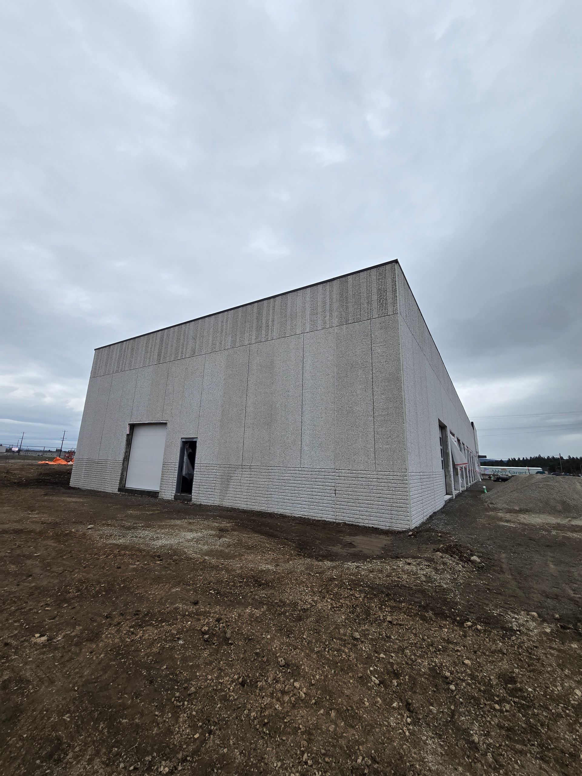 A large, white, 3D-printed concrete warehouse under a cloudy sky, situated on an unpaved, dirt construction site.