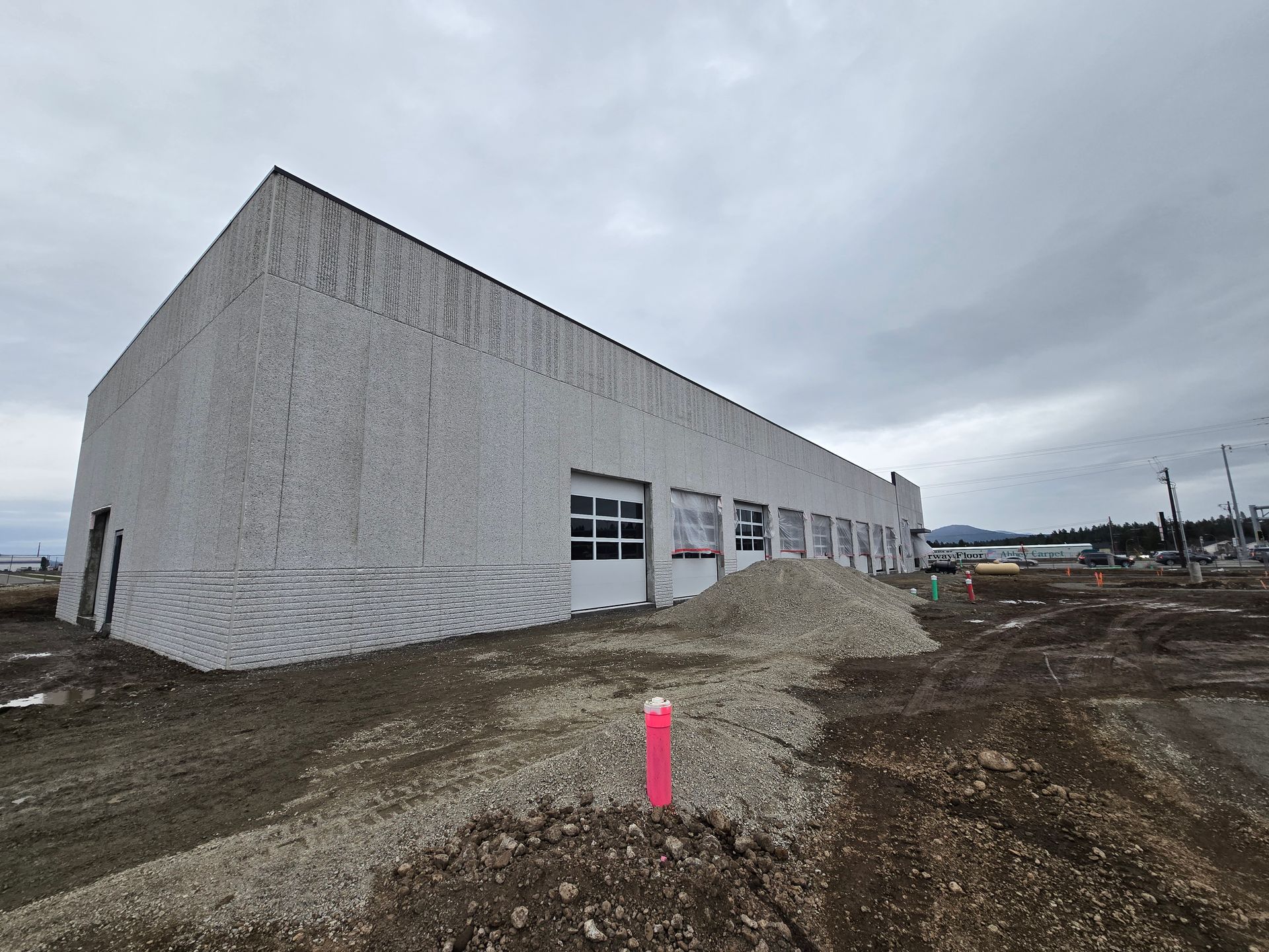 A long, windowless warehouse building under construction with a gravel yard and a pink survey marker in the foreground.