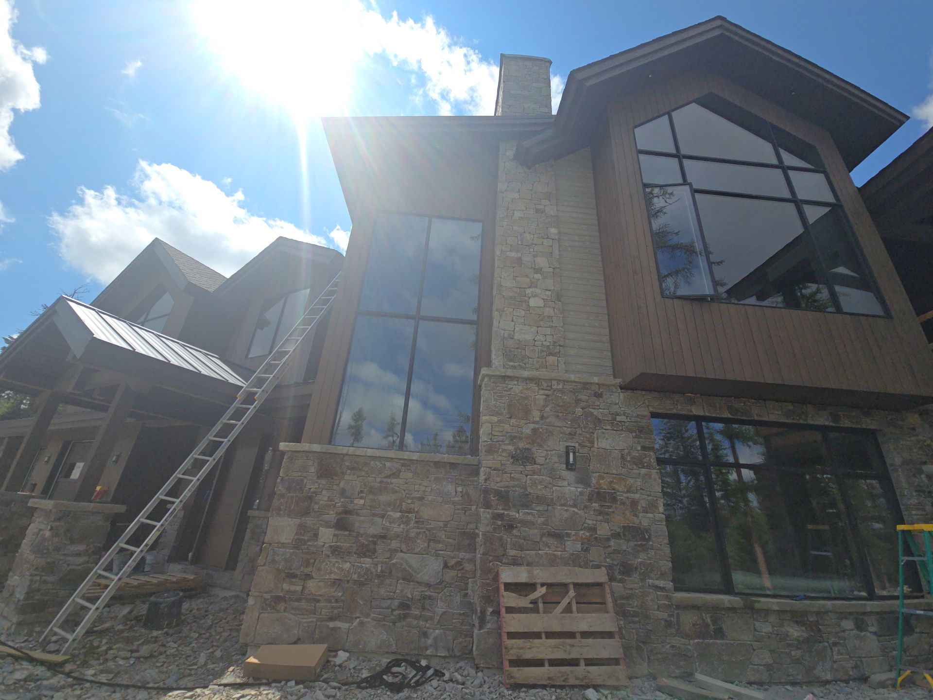 An unfinished house with stone veneer, brown vertical siding, and large windows under a bright, sunny sky.