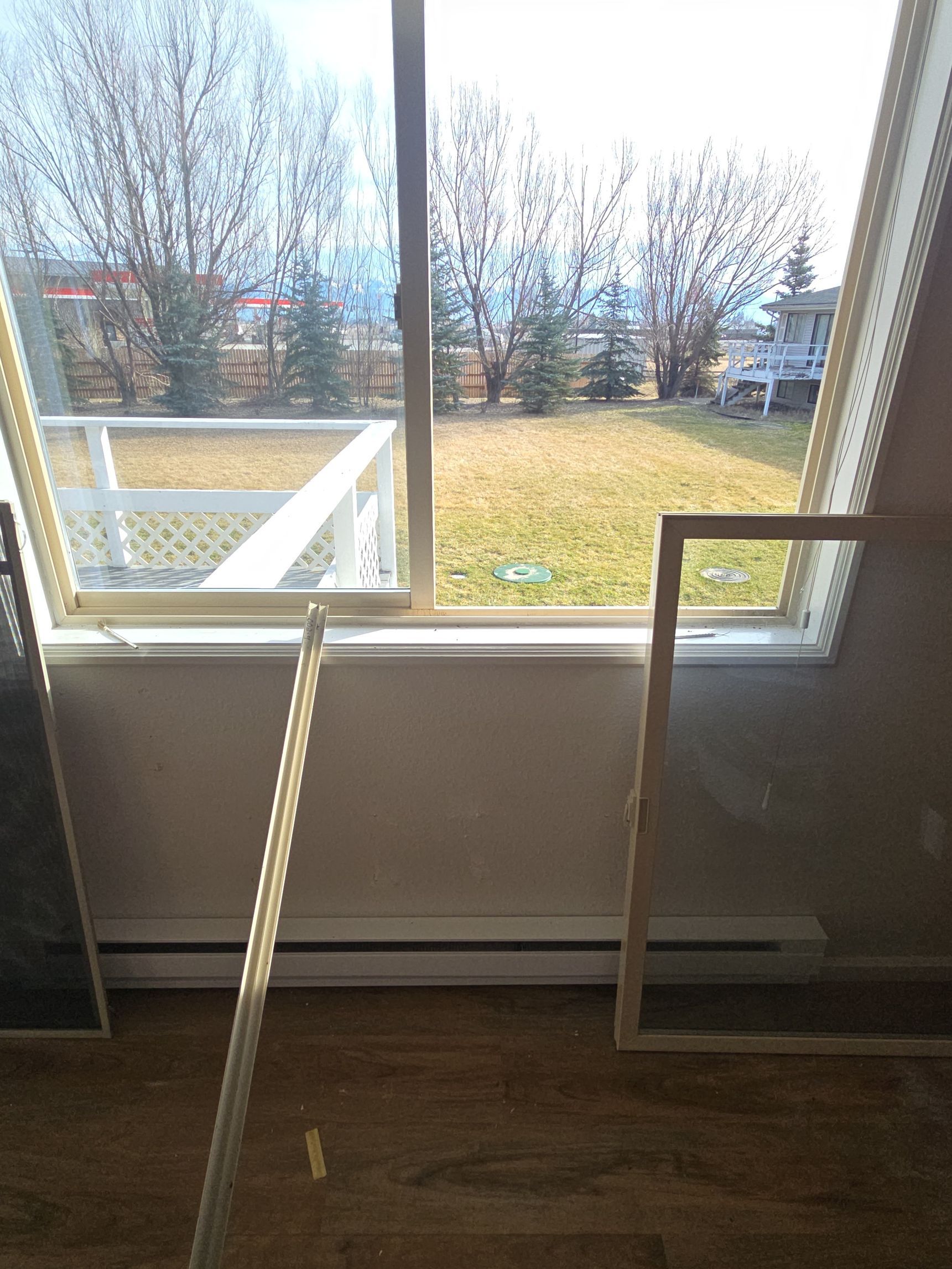 A sliding glass door with detached screens leaning against the wall, overlooking a yard with trees and a white fence.