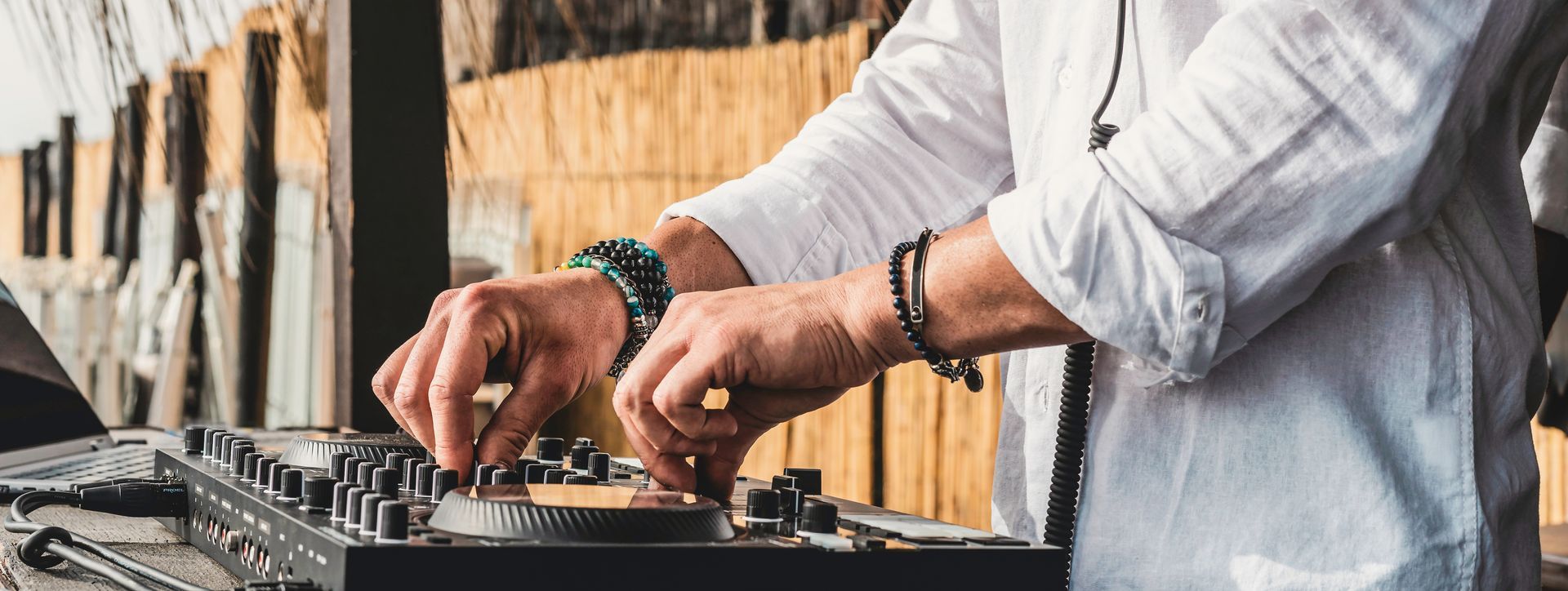 Hands of a DJ operating a mixing console. He wears a white shirt and bracelets.