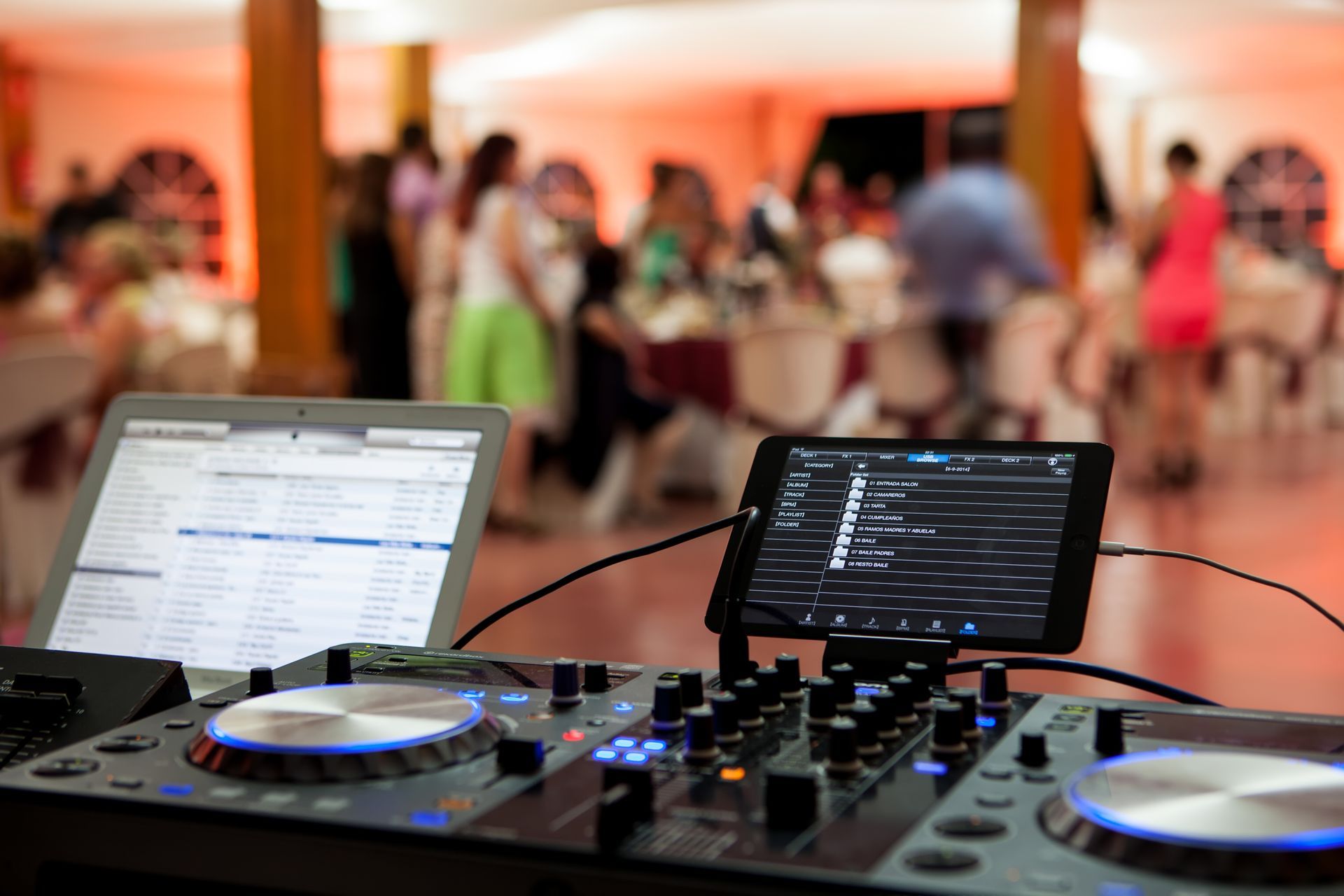 DJ setup at a party with a laptop, tablet, and mixer. Blurred background of people dancing and socializing.