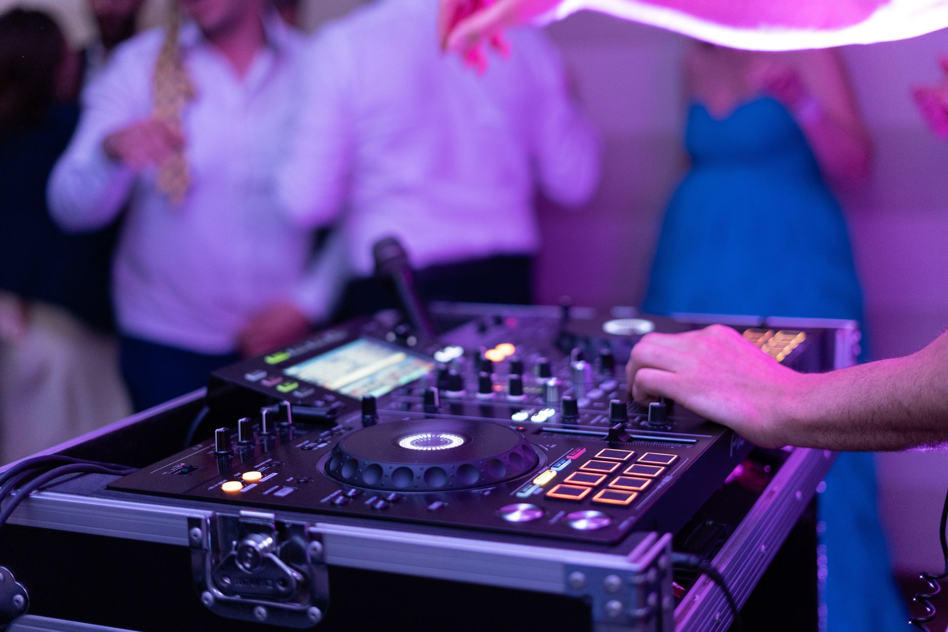 DJ using soundboard with purple and pink lighting, people dancing in the background.