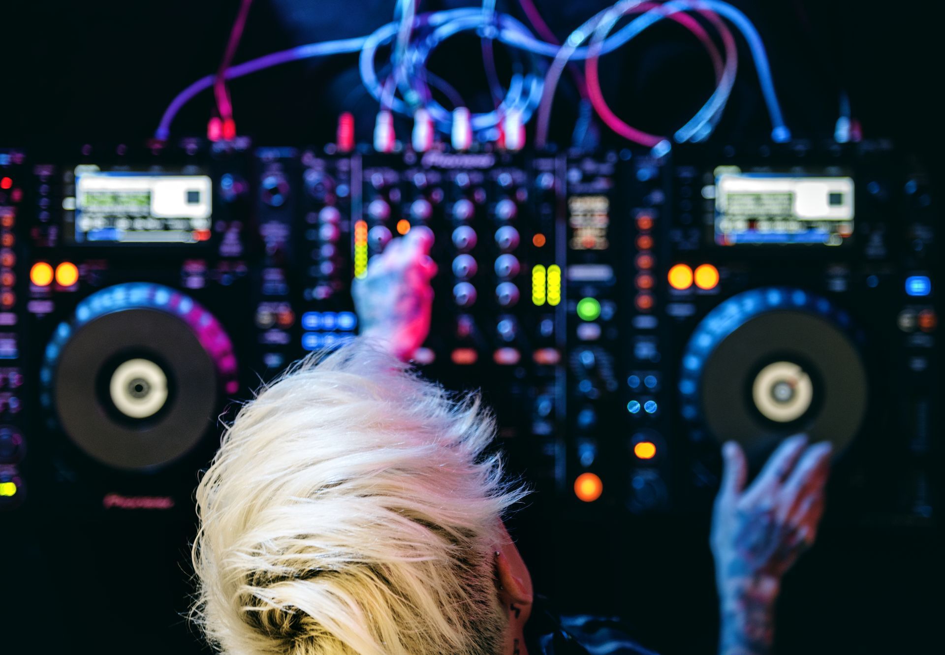 DJ with light blonde hair, mixing music on a brightly lit console with colorful lights in a dark setting.