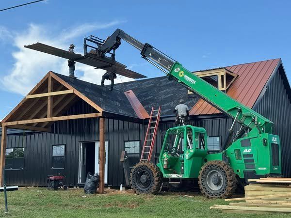 Workers using a green telehandler to install roofing on a black house with a brown metal roof.