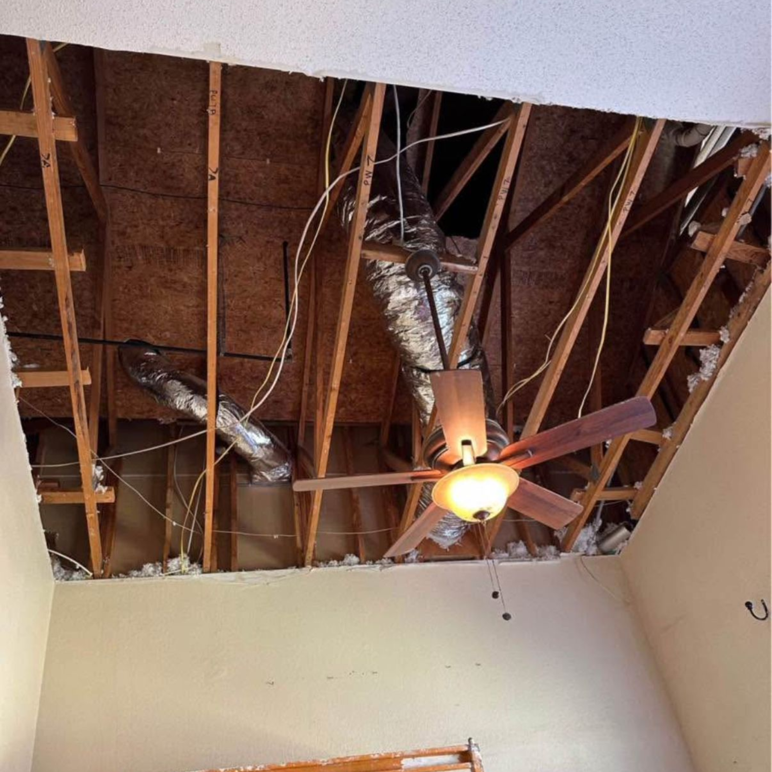 Ceiling partially collapsed, revealing wooden beams, ductwork, and wiring. A ceiling fan hangs below.