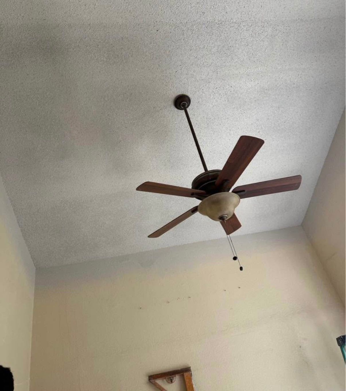 Ceiling fan with five brown blades on a textured ceiling in a room with tan walls.