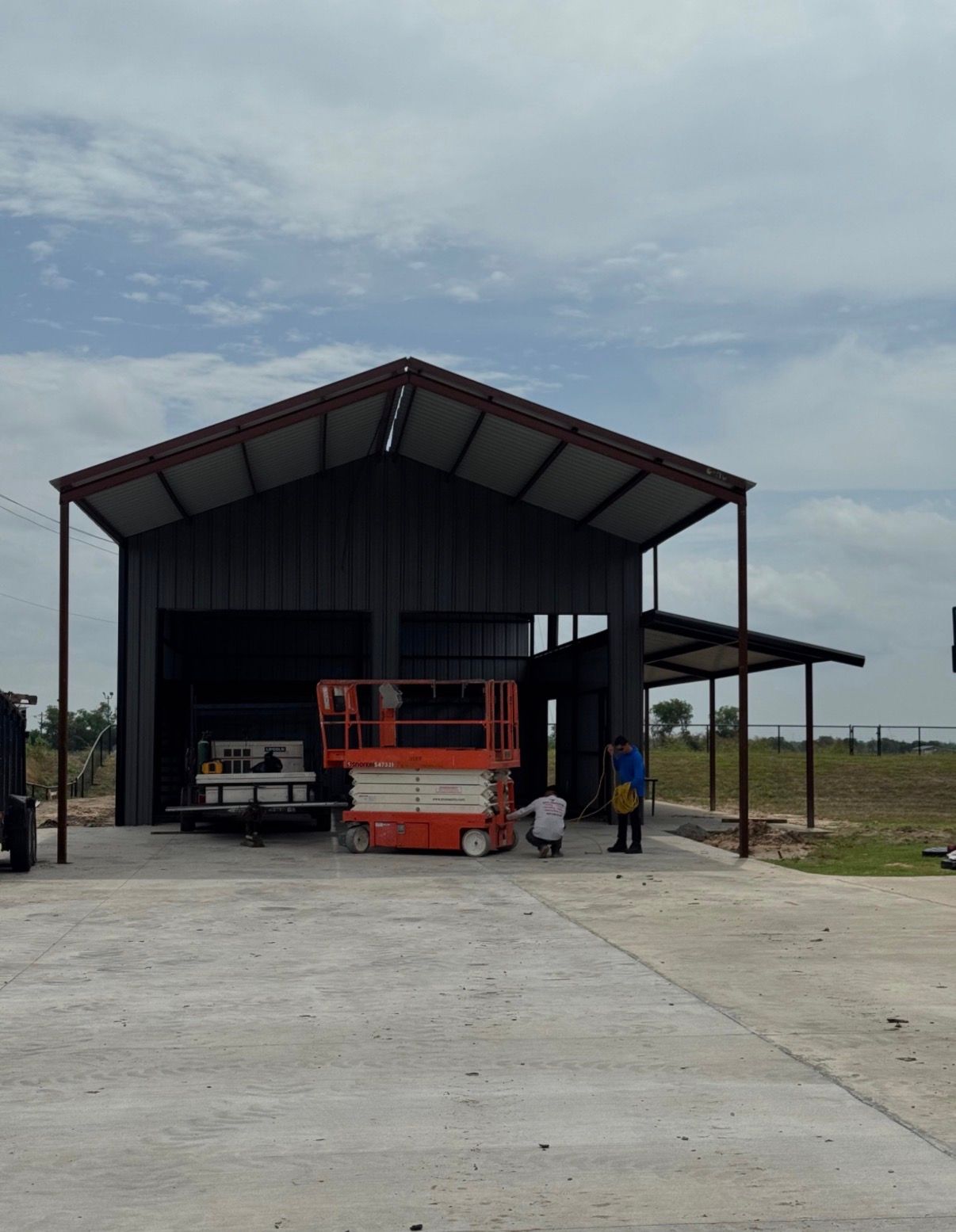 Metal building with lift and two people, open bay, concrete apron, cloudy sky.