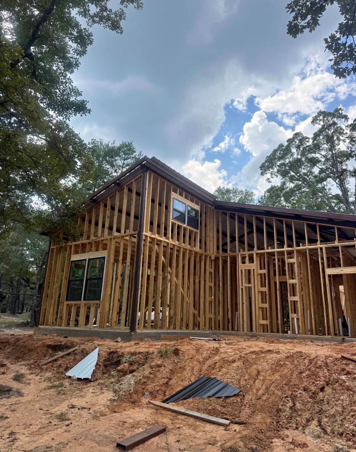 Wood frame of a two-story house under construction. Exterior walls, windows visible, set in a wooded area. Cloudy sky.