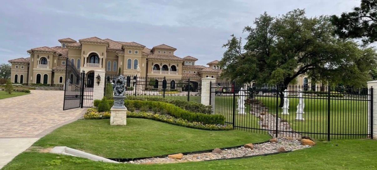 A large beige mansion with a black wrought iron fence and gate, a driveway, and a tree in the yard.
