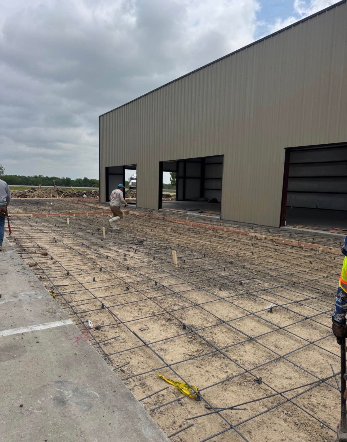 Construction site; building with open bays, workers, rebar grid on ground, cloudy sky.