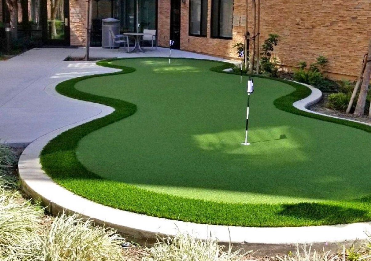 A putting green with three holes in a backyard. Green turf, white flags, and a concrete border.