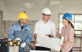 Three construction workers discussing blueprints in a building. Two wear hard hats, one is pointing.