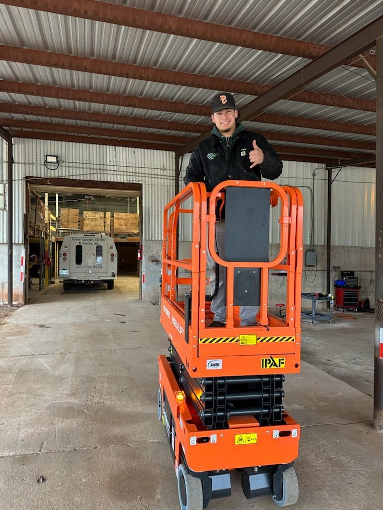 A person in a black jacket and baseball cap smiles and gives a thumbs-up while standing on an orange scissor lift.