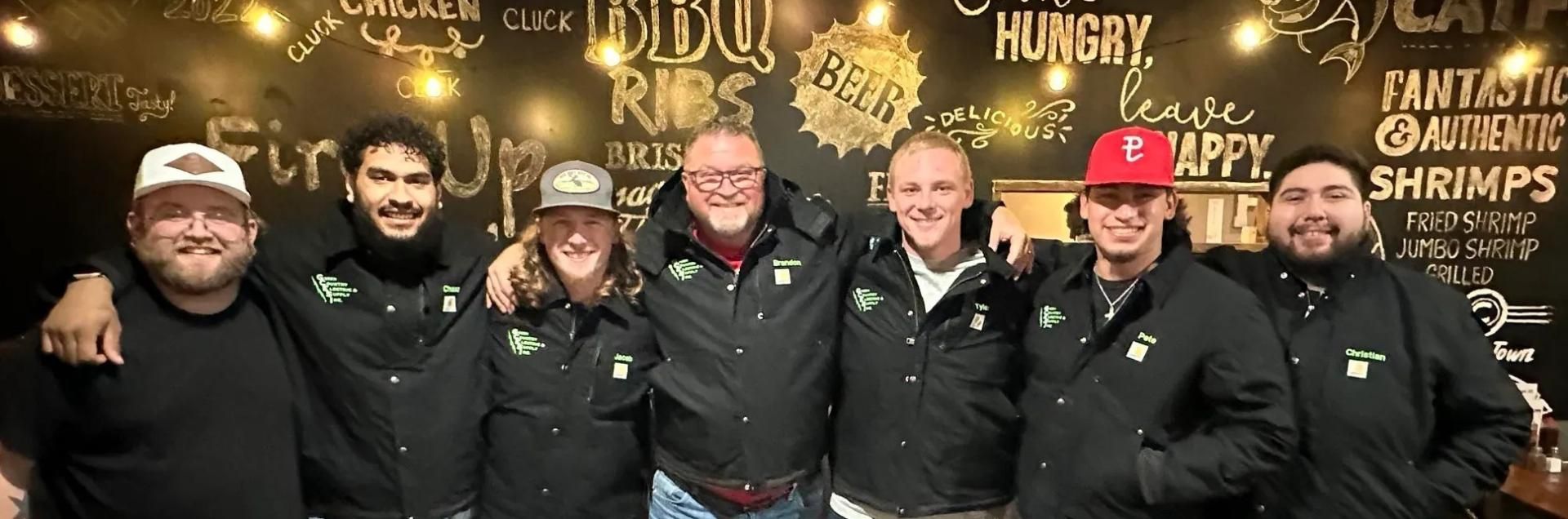A group of seven people smiling in matching black jackets, standing in front of a wall with stylized restaurant text.