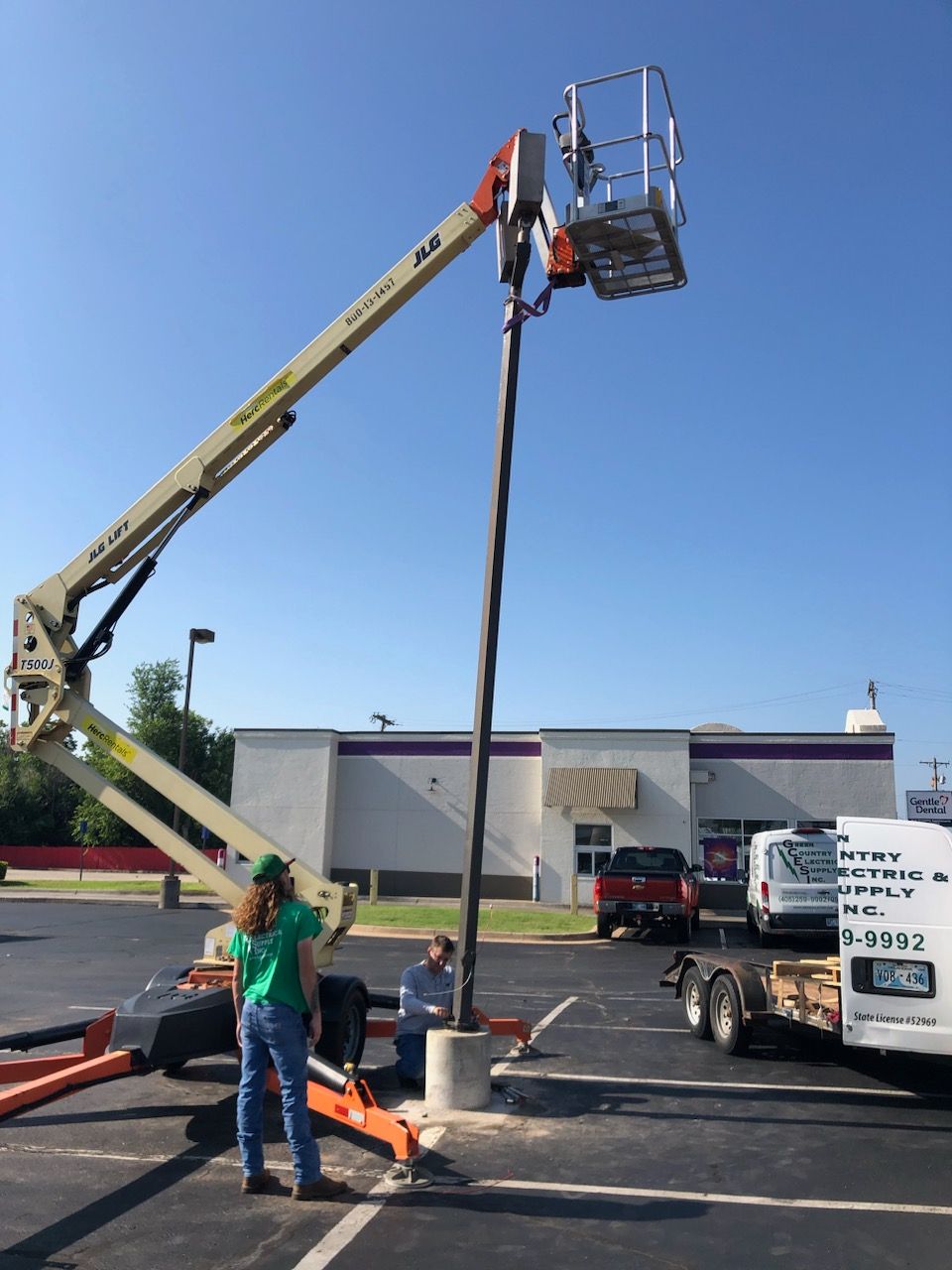 Two people work on a tall parking lot light pole using an elevated aerial lift truck in a paved lot.