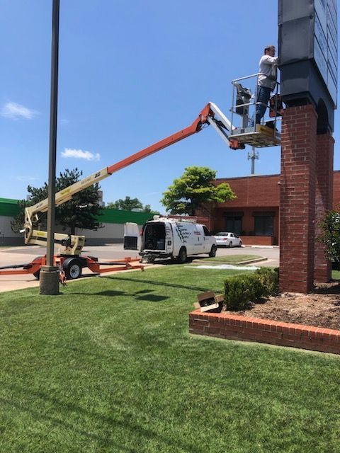 A technician in a bucket lift services a large, brick-supported sign outdoors on a sunny day next to a parked utility van.