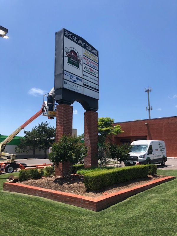 A worker in an aerial lift services a Broadway Square shopping center monument sign set in a landscaped brick planter.