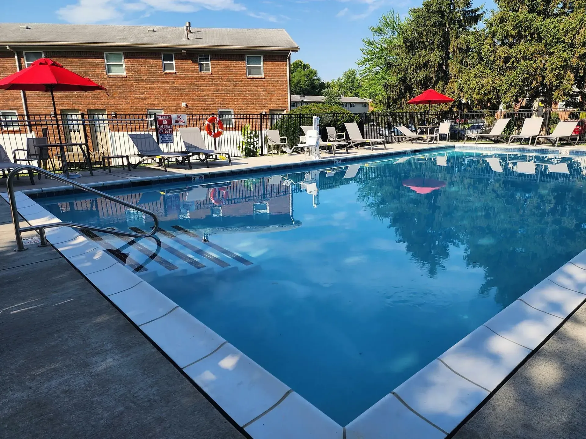 Swimming pool with lounge chairs, two red umbrellas, and a brick building on a sunny day.