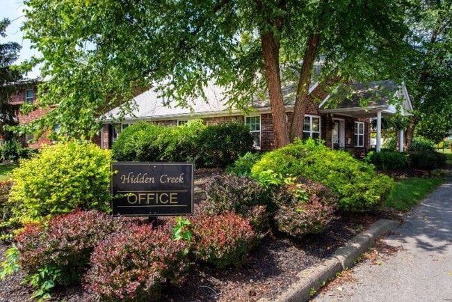 Hidden Creek Office sign in front of a brick building surrounded by green and red bushes and trees.