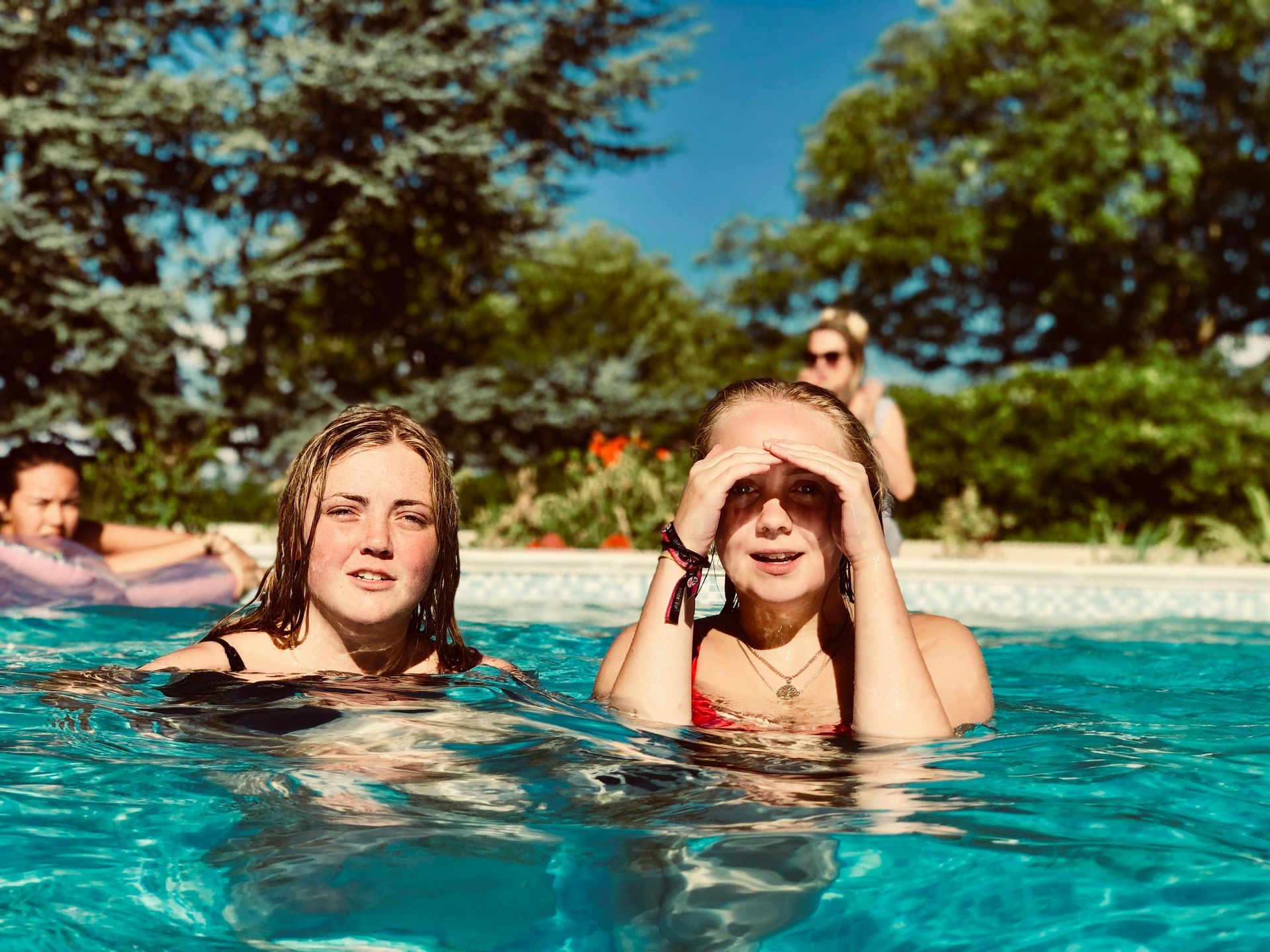 Two girls in a pool, one shielding eyes, other looks at the camera. Lush trees in the background, sunny day.