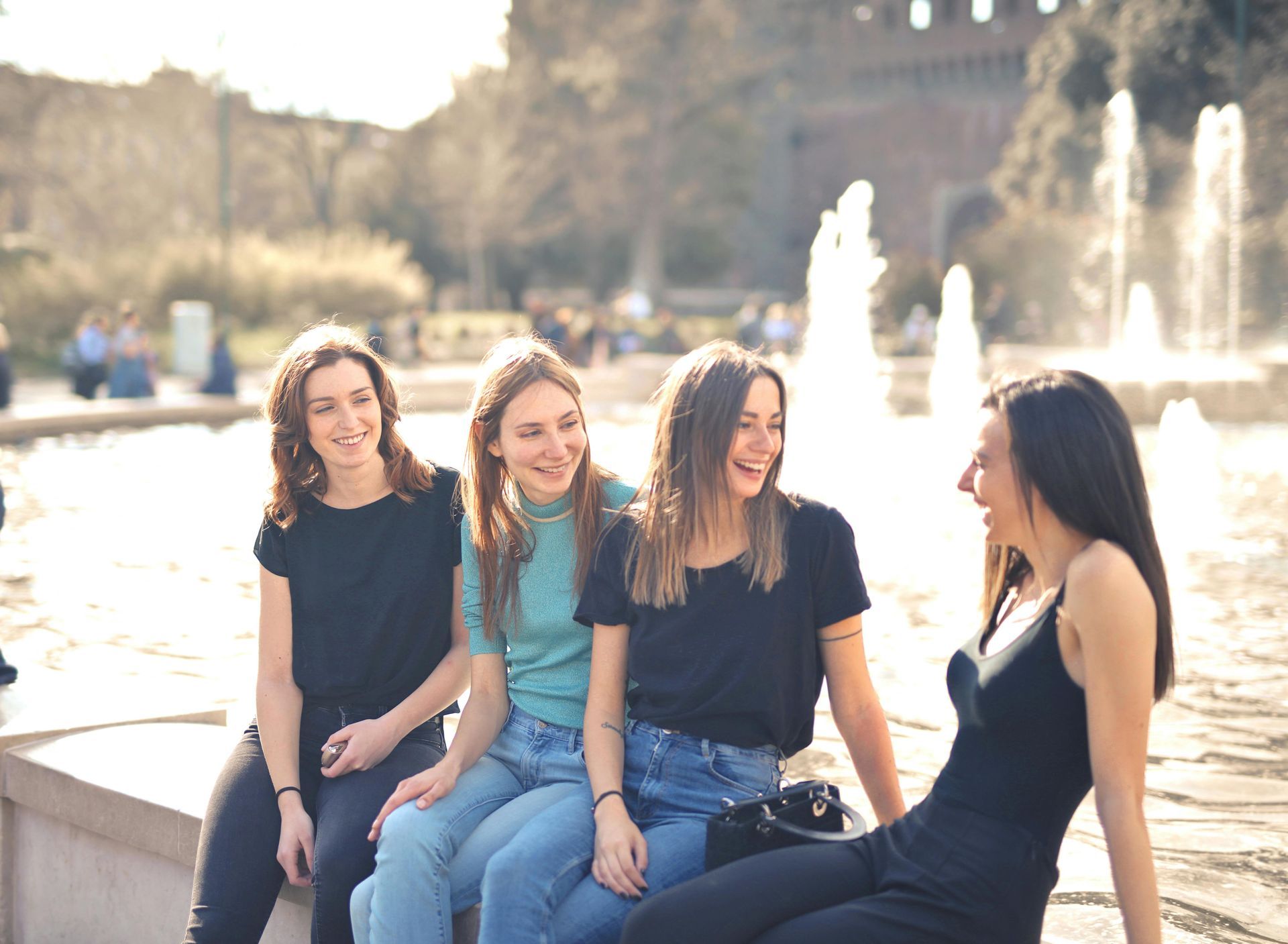 Four women laugh together outdoors near a fountain.