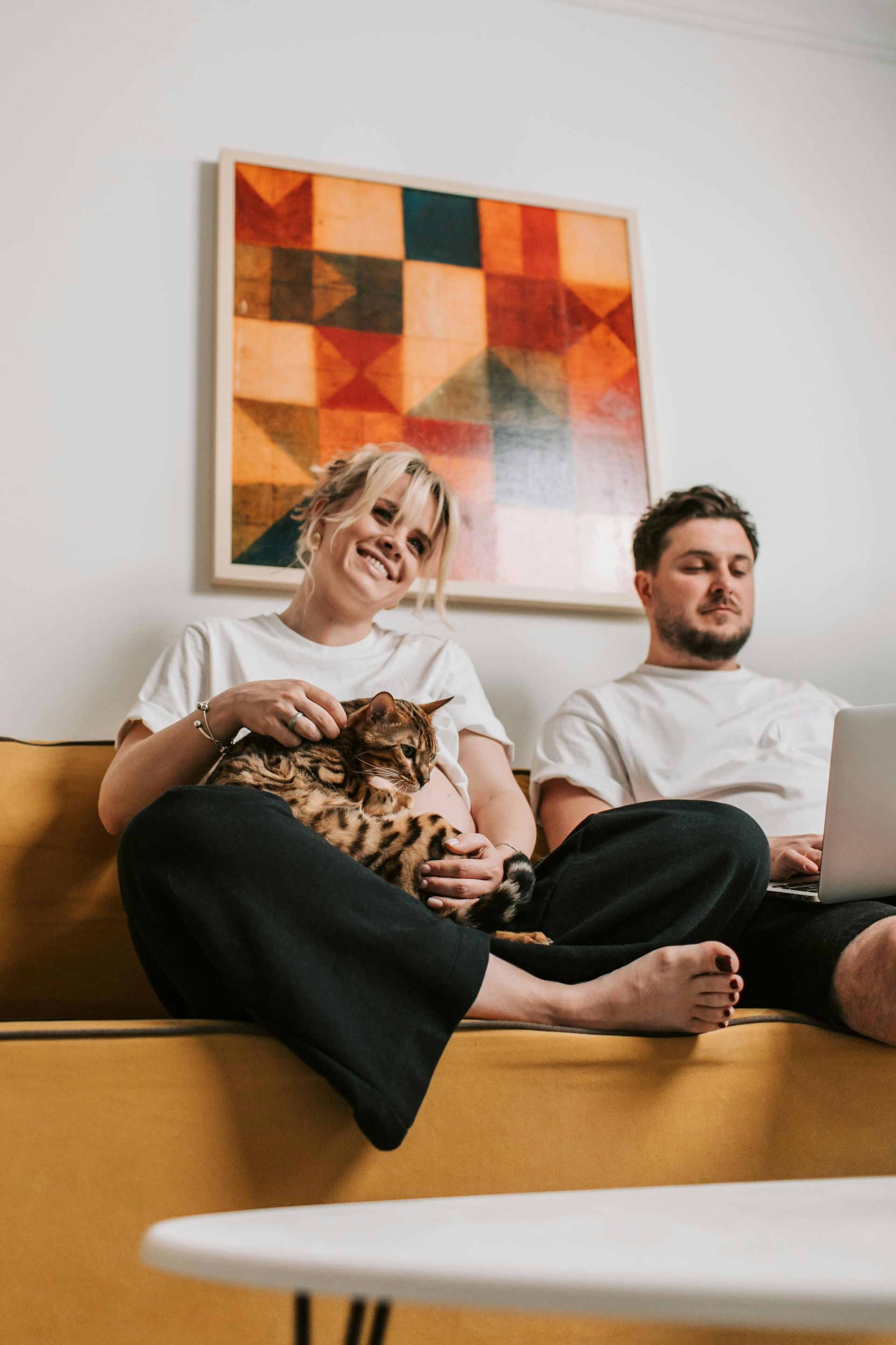 Woman smiles, holding a cat, next to a man using a laptop, sitting on a yellow couch. Modern art above them.