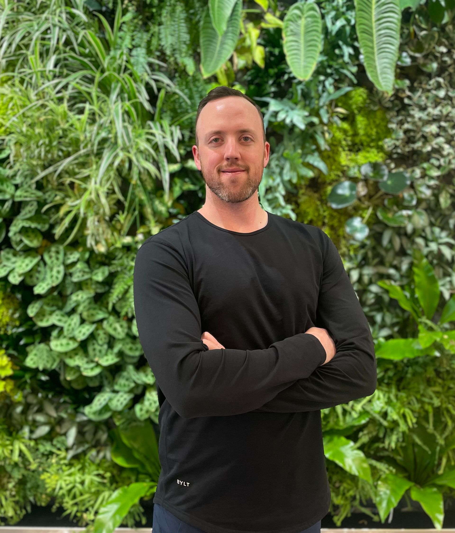A man in a black shirt is standing in front of a wall of plants.