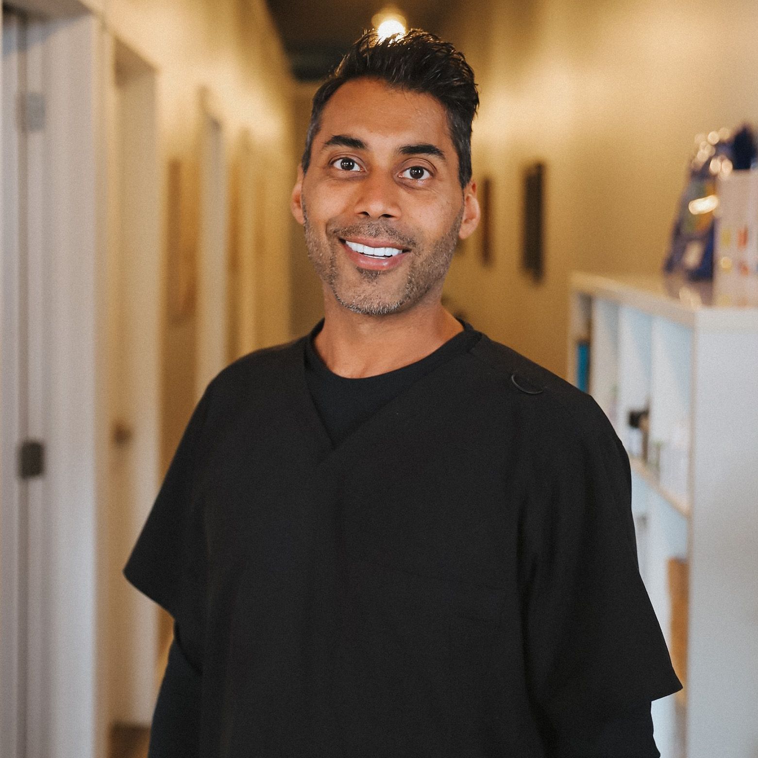 A man in a black shirt is smiling for the camera in a hallway.