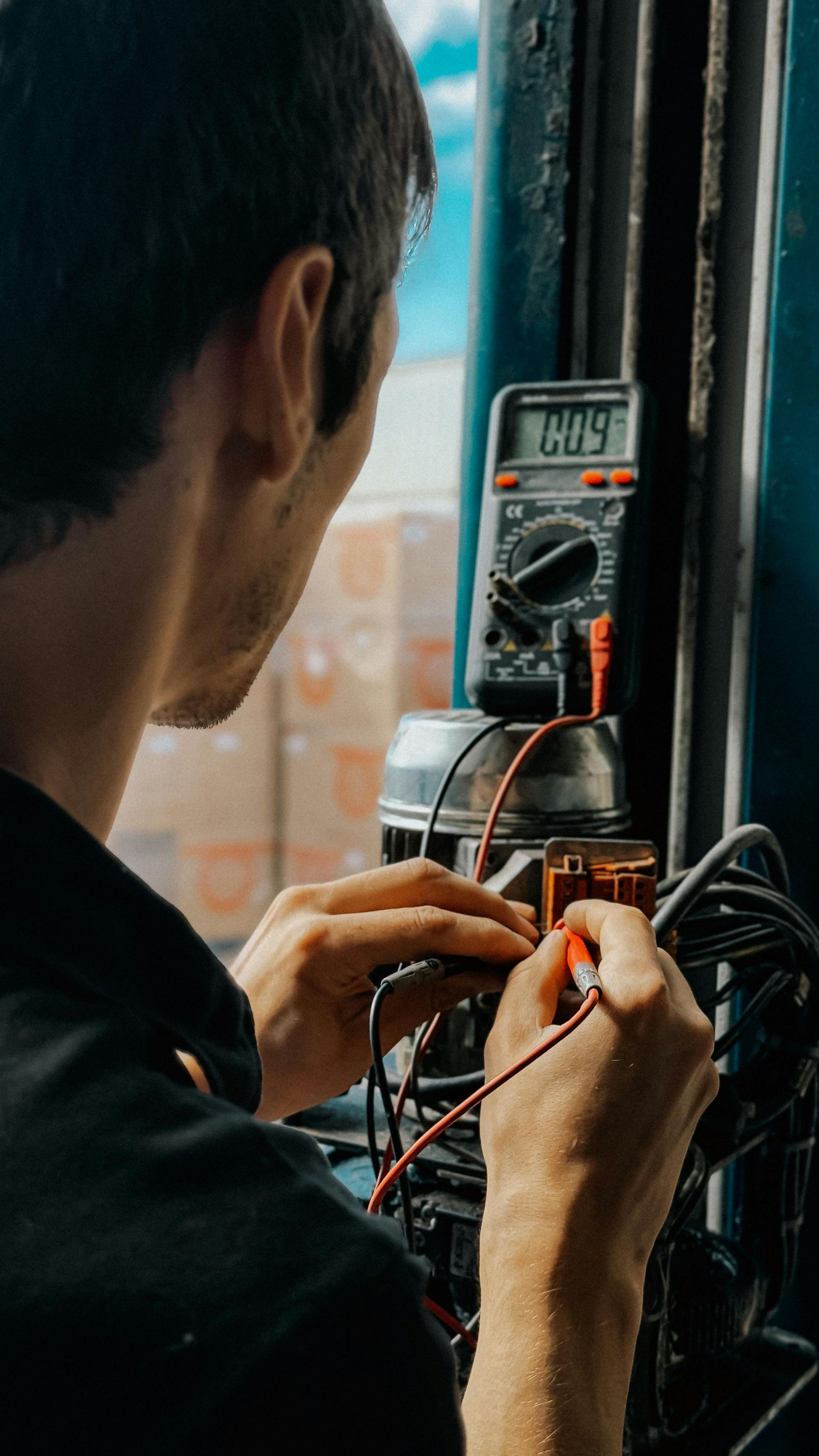 Person testing electrical wiring with a multimeter at a window, wearing a black jacket.