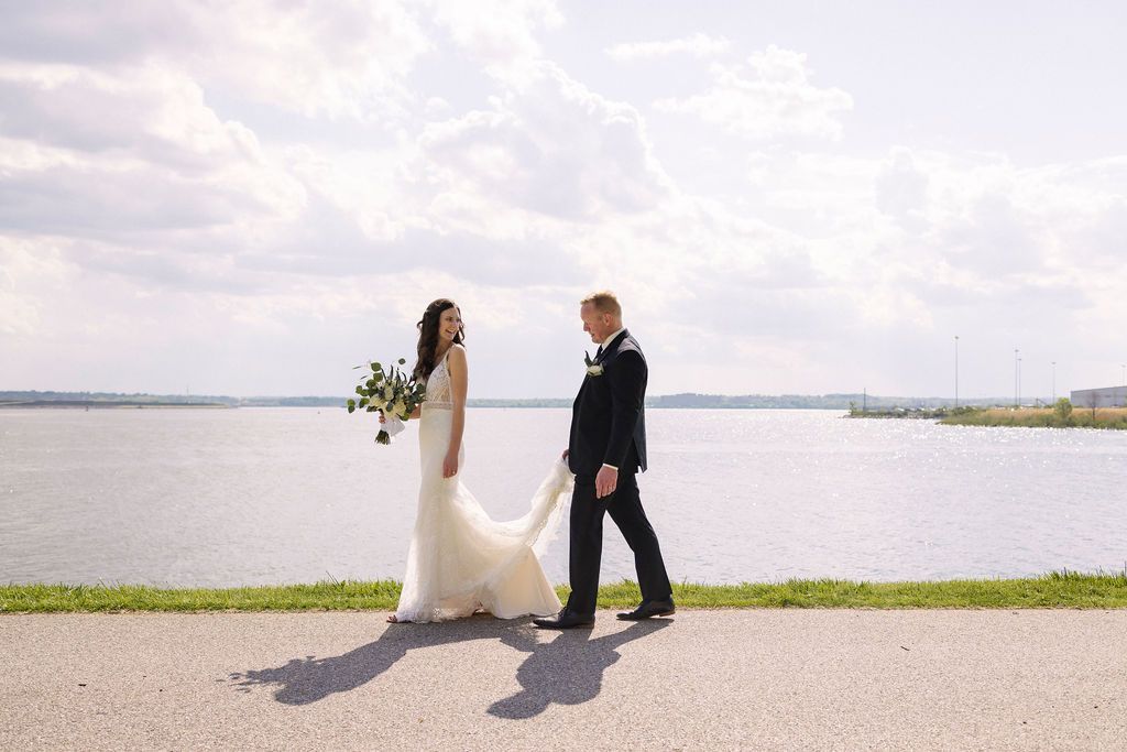 Bride looks back at groom as he steps, holding her dress train. Waterscape backdrop.