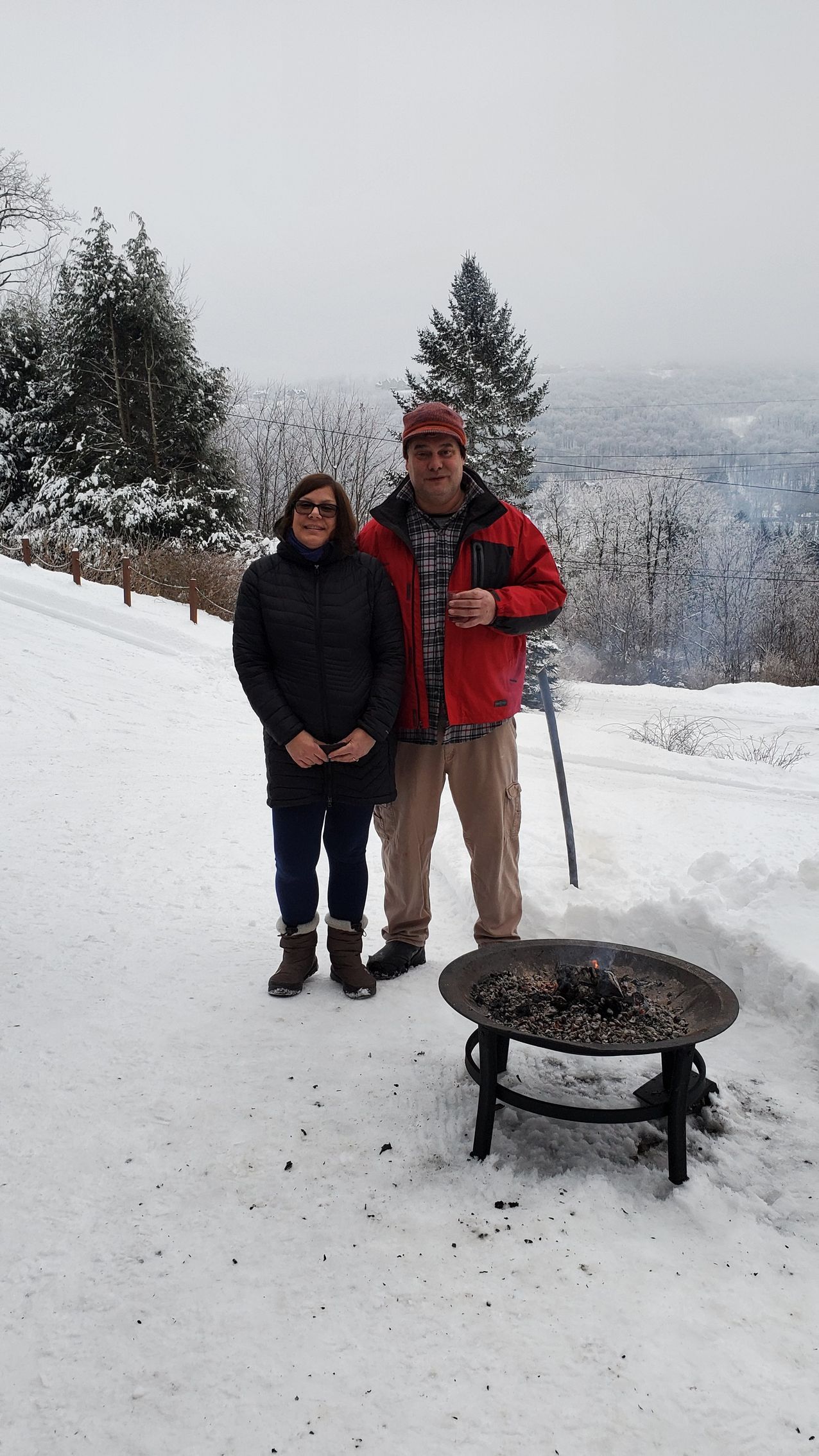 Couple stands in snow next to a fire pit, overlooking a snowy valley. Man holds a cup, and wears a red jacket.