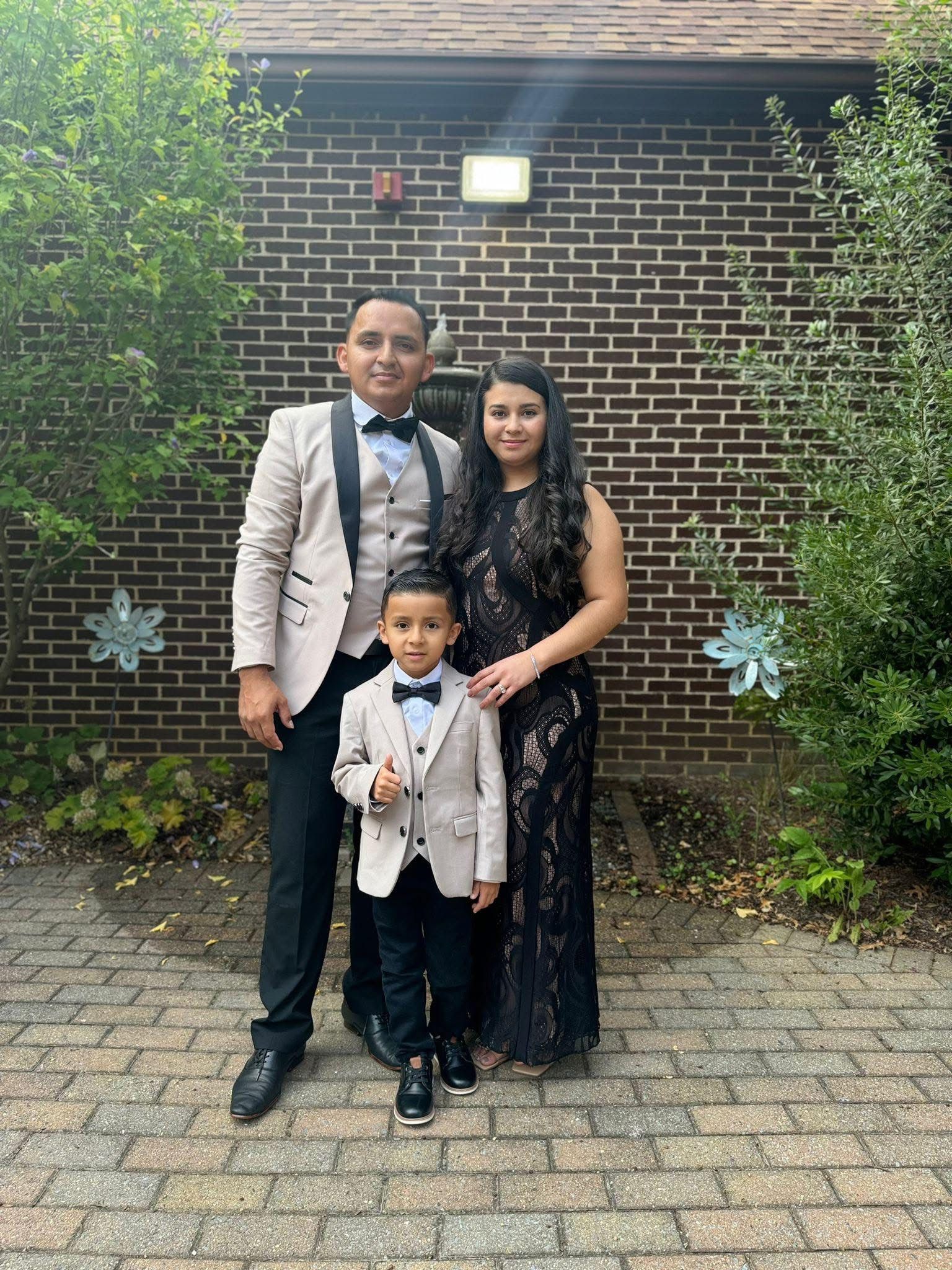 Family of three dressed up in front of a brick building. Man and boy wear tan jackets, woman wears black gown.