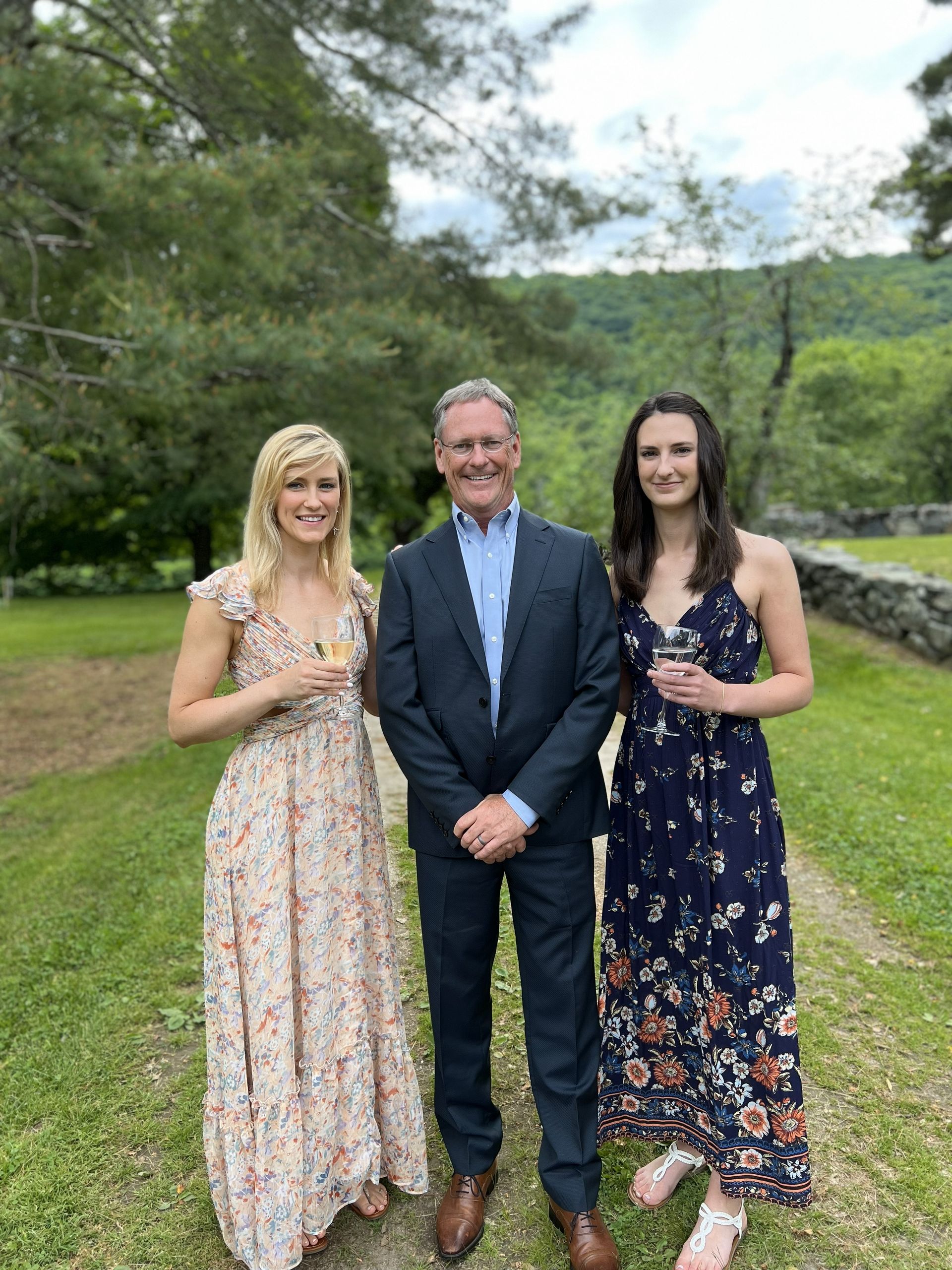 Three people smiling outdoors on a path: a man in a suit between two women in floral dresses, holding drinks.