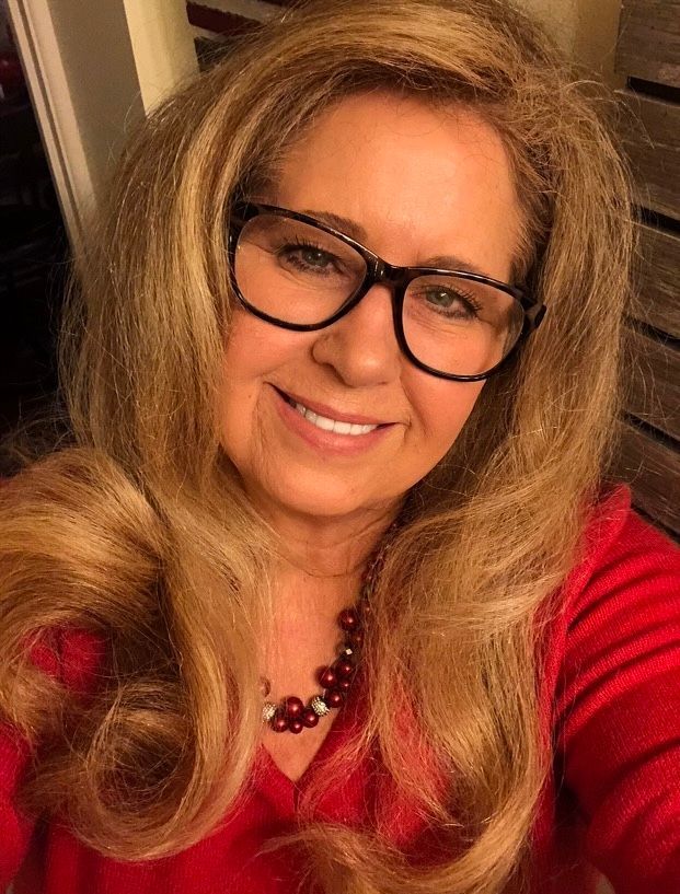 Woman with long blonde hair, glasses, and red top smiles at the camera; a red necklace is visible.