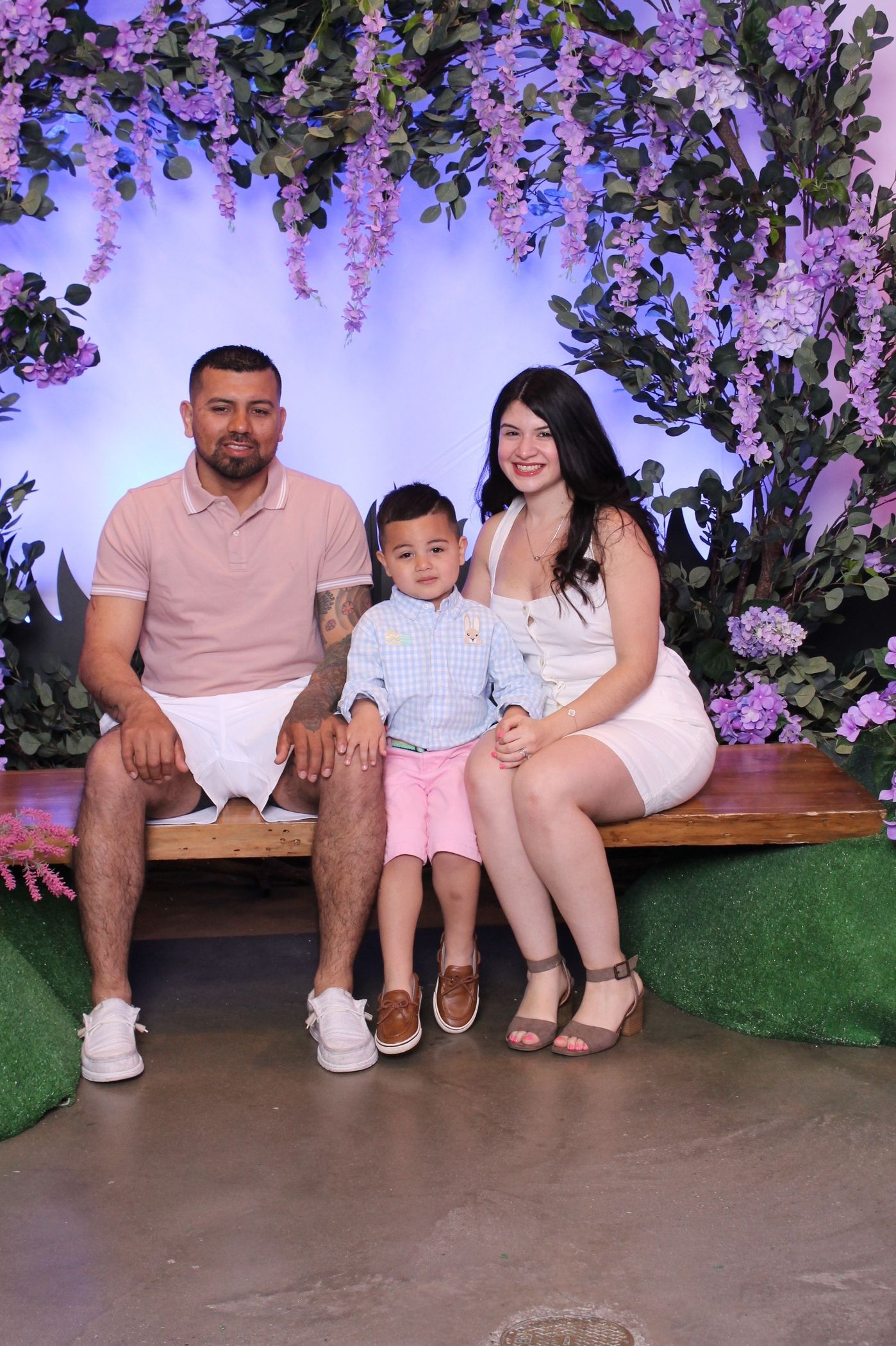 Family posing on a bench in front of purple flowers. Man in pink shirt, woman in white dress, child in blue.