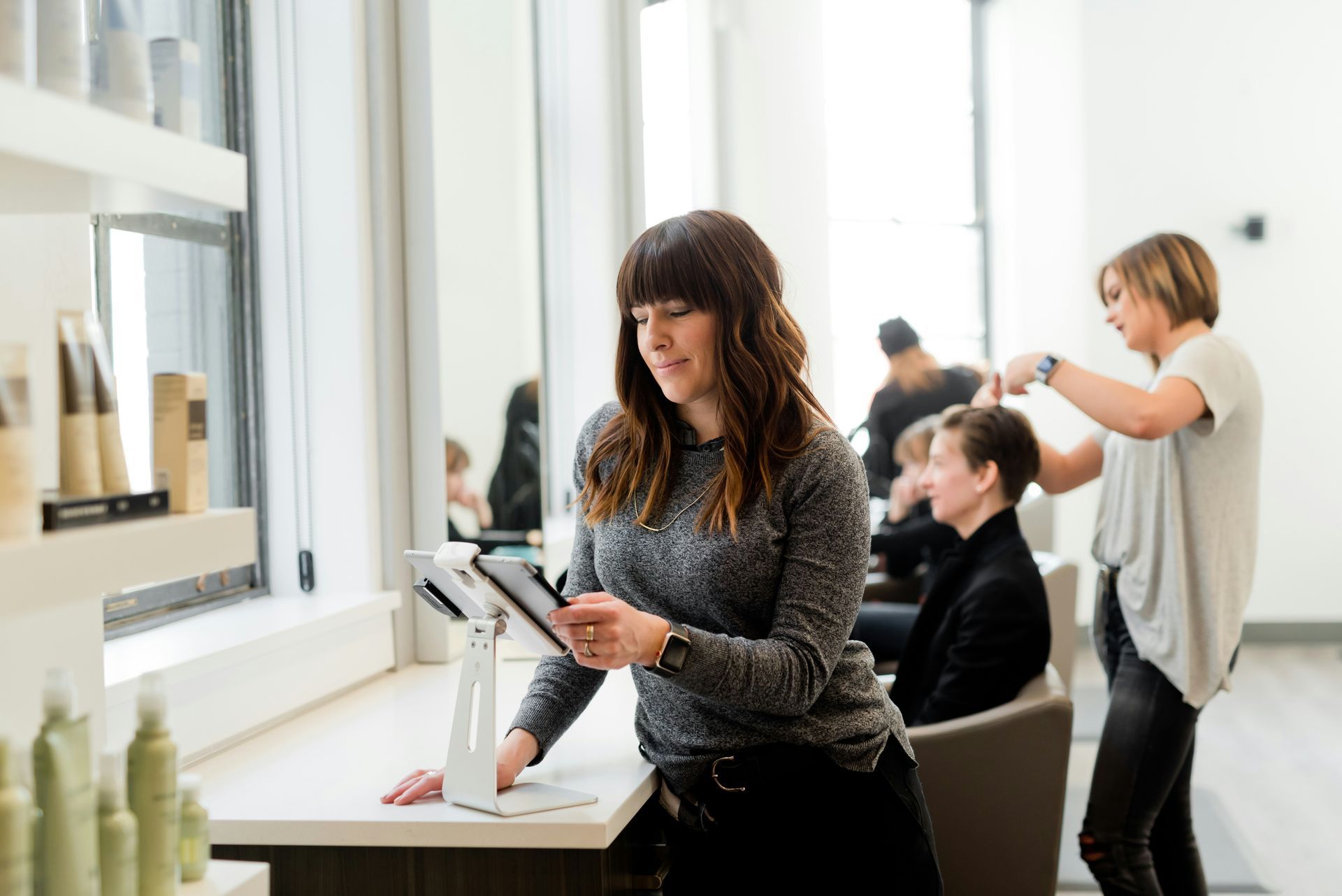 Woman using tablet at a salon counter; clients getting haircuts.