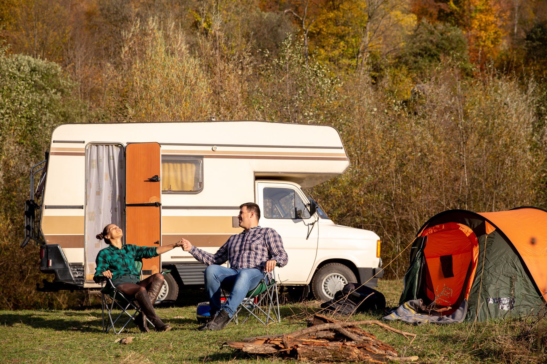 Couple camping in front of a camper and tent in a wooded area on a sunny day.