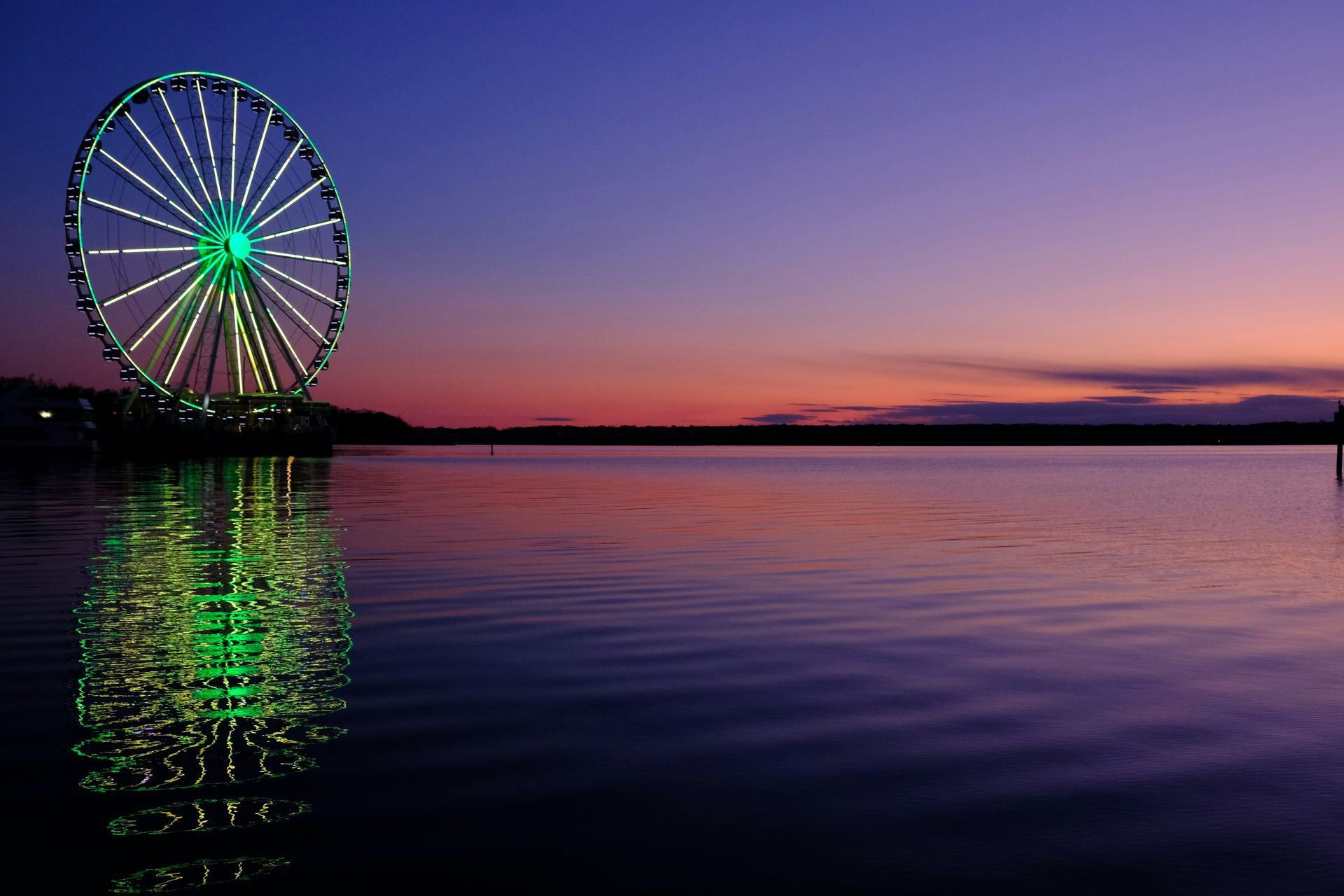 A lit Ferris wheel reflecting in calm water at sunset.