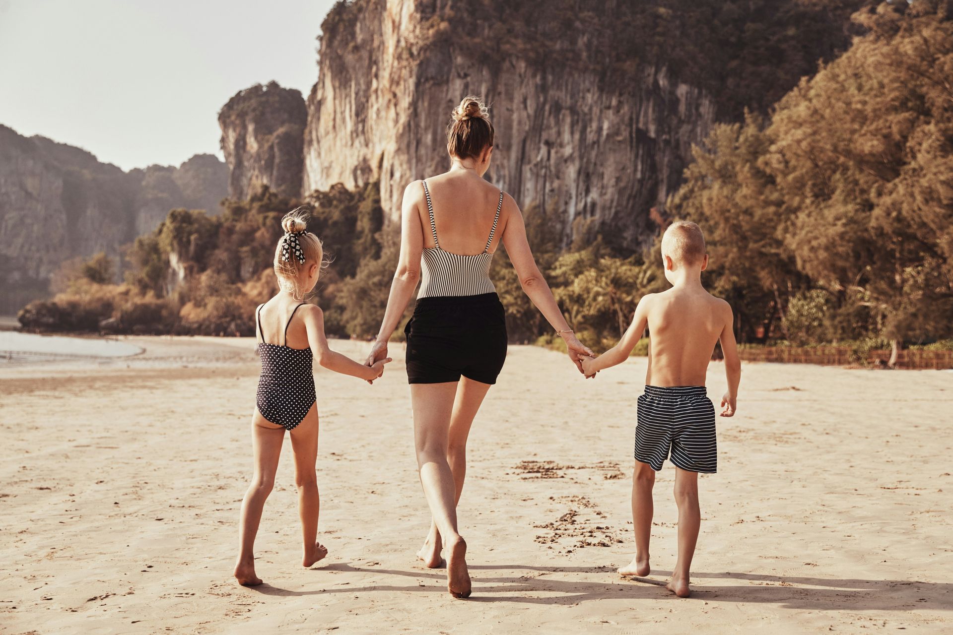 Woman holding hands with two children on a sandy beach, mountains in the background.