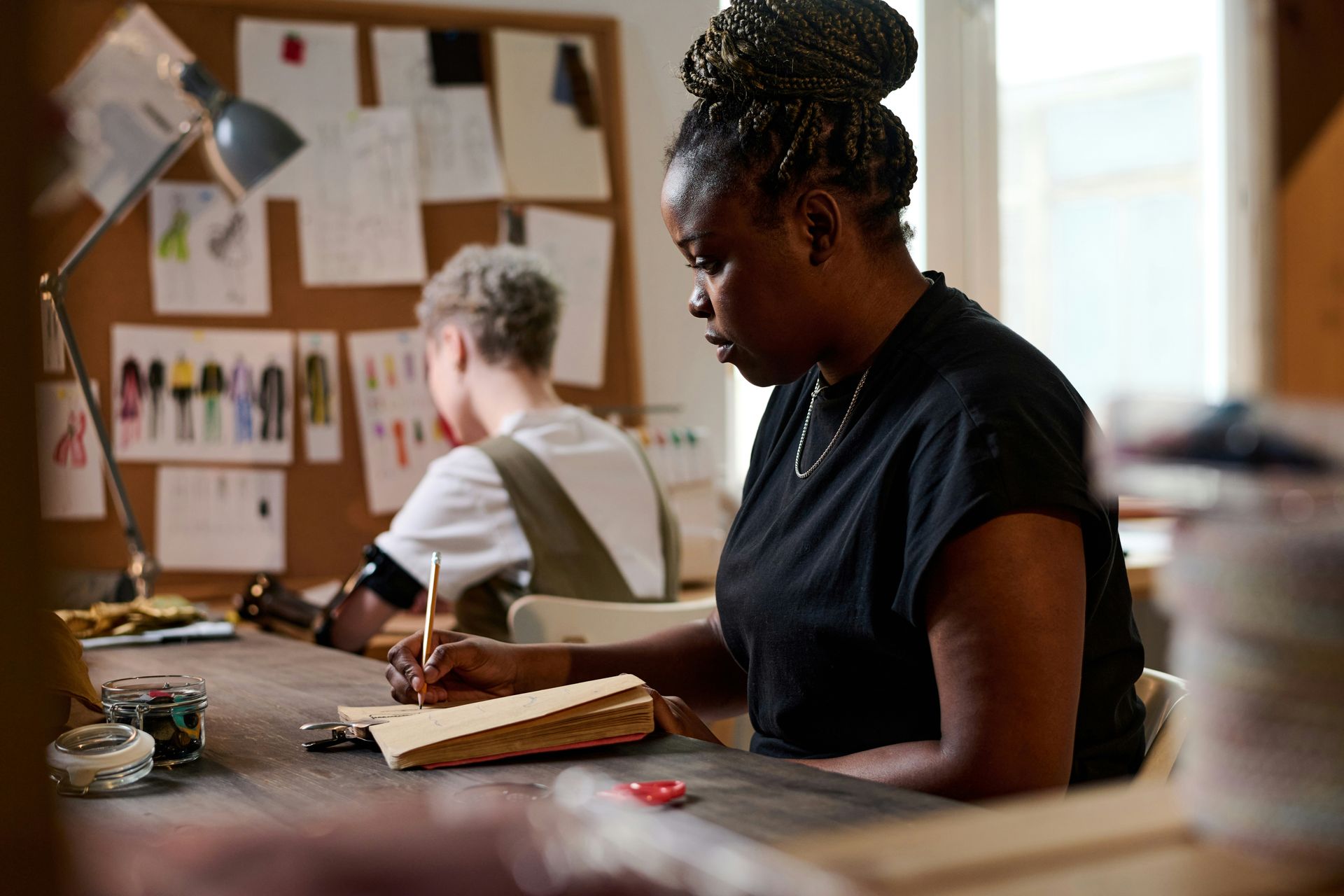 Woman writing in a notebook at a desk, fashion design studio. A second person is in the background.