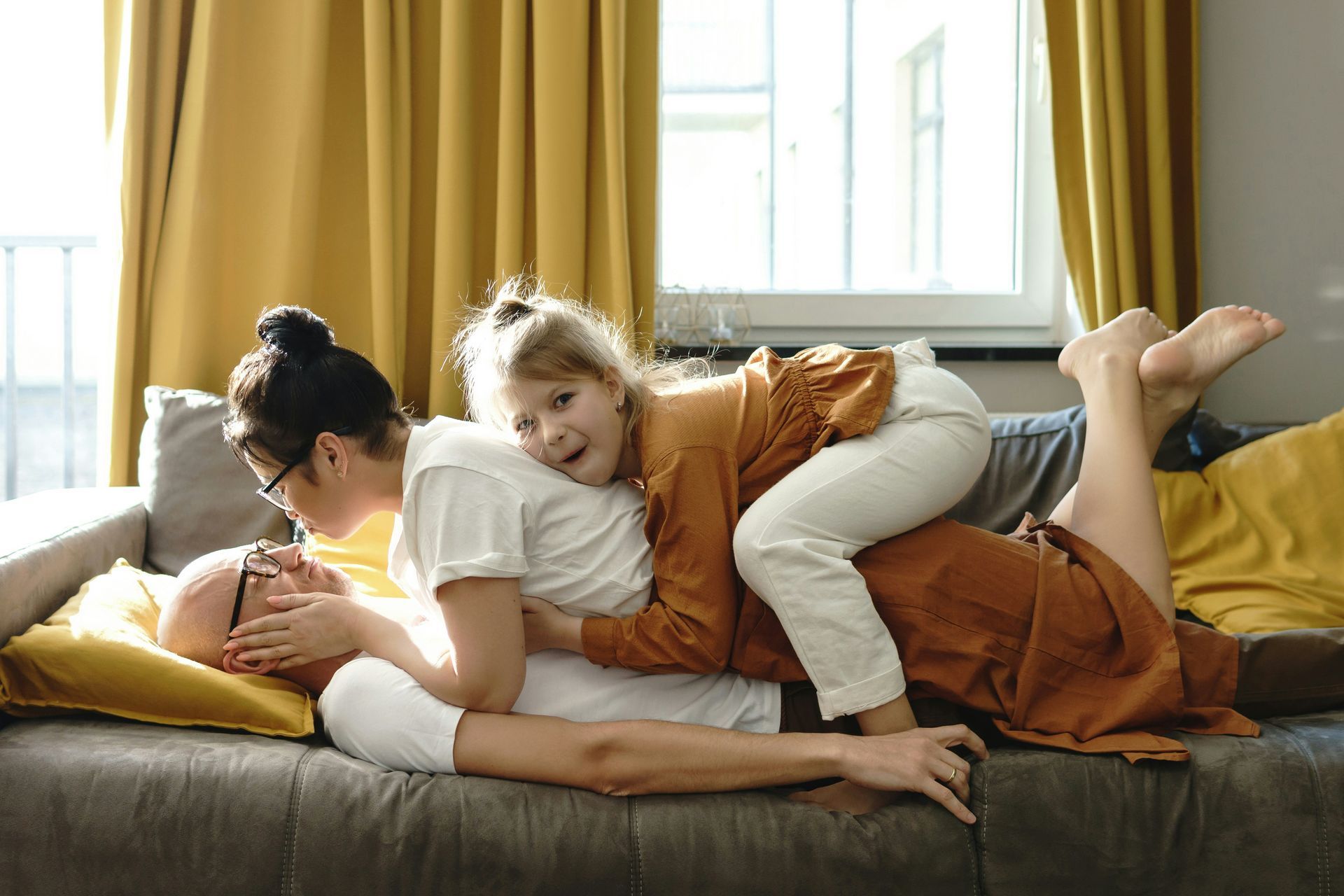 Family playfully piled on a couch: a child on the father's back while the mother covers his eyes, all smiling.