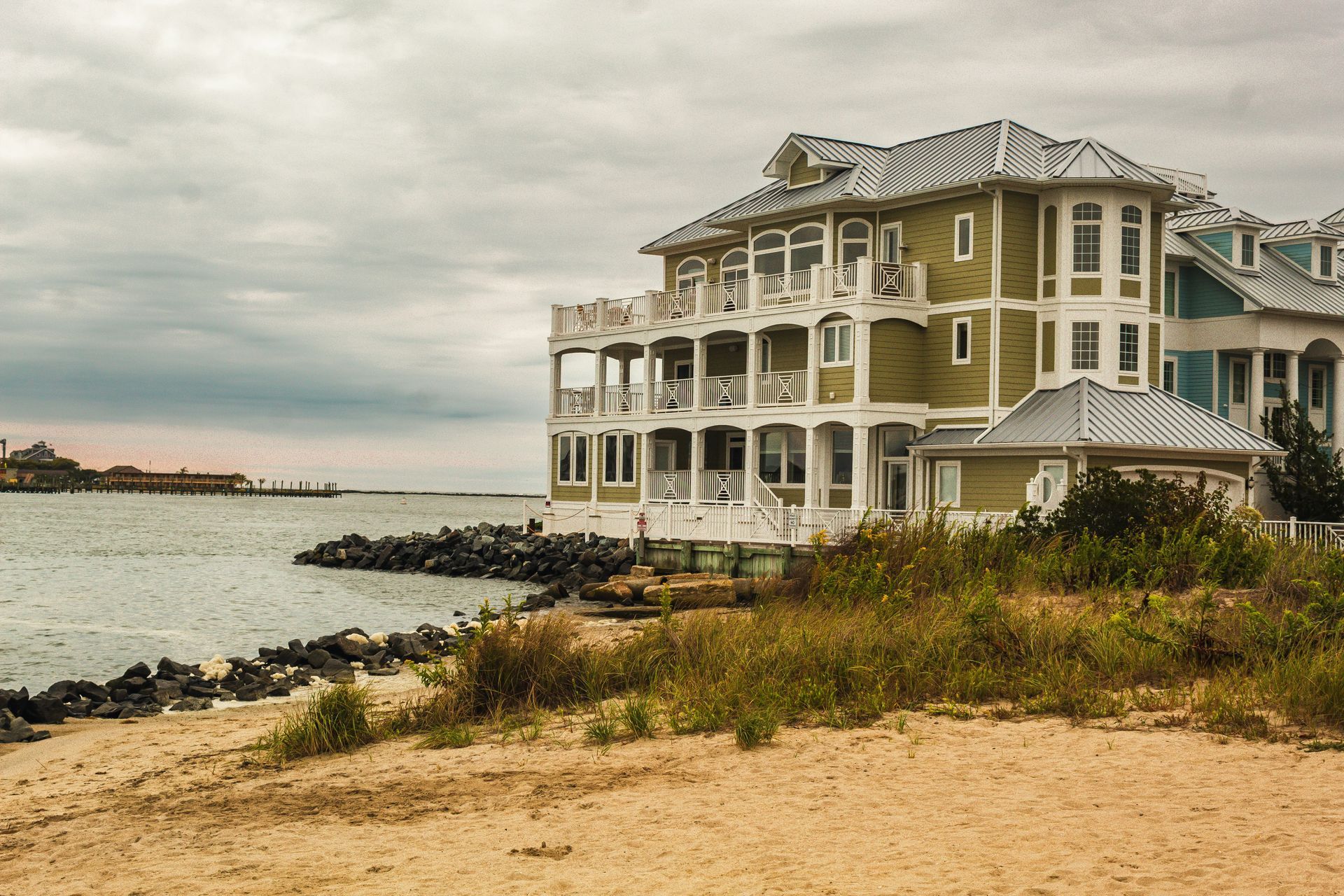 Large multi-story house on a sandy beach, green and blue exterior, overlooking water. Overcast sky.
